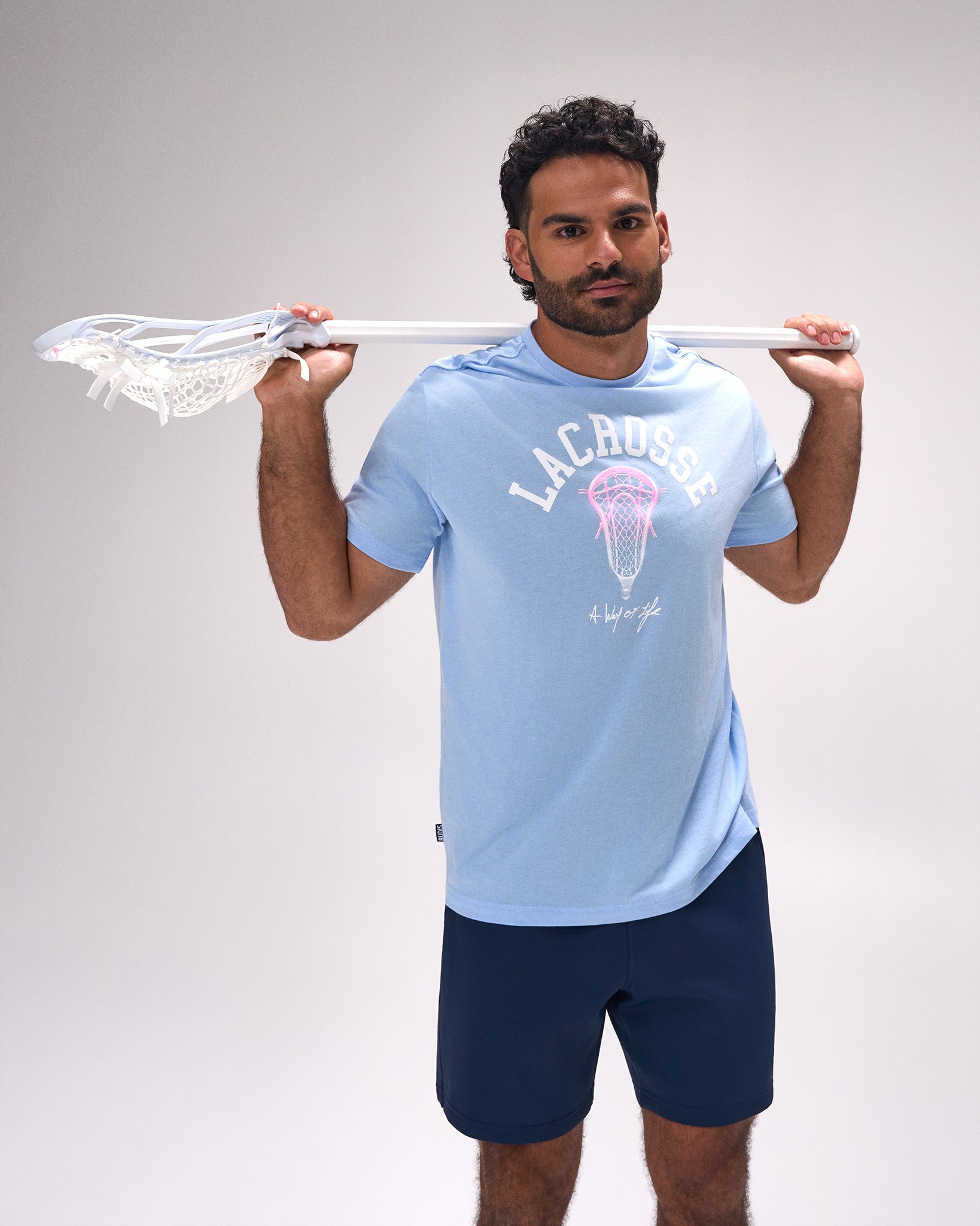 Male athlete wearing the RUDIS LAX Motto Elite Super Soft T-Shirt in Light Blue featuring bold "Lacrosse" graphic with lacrosse head artwork while holding a lacrosse stick.