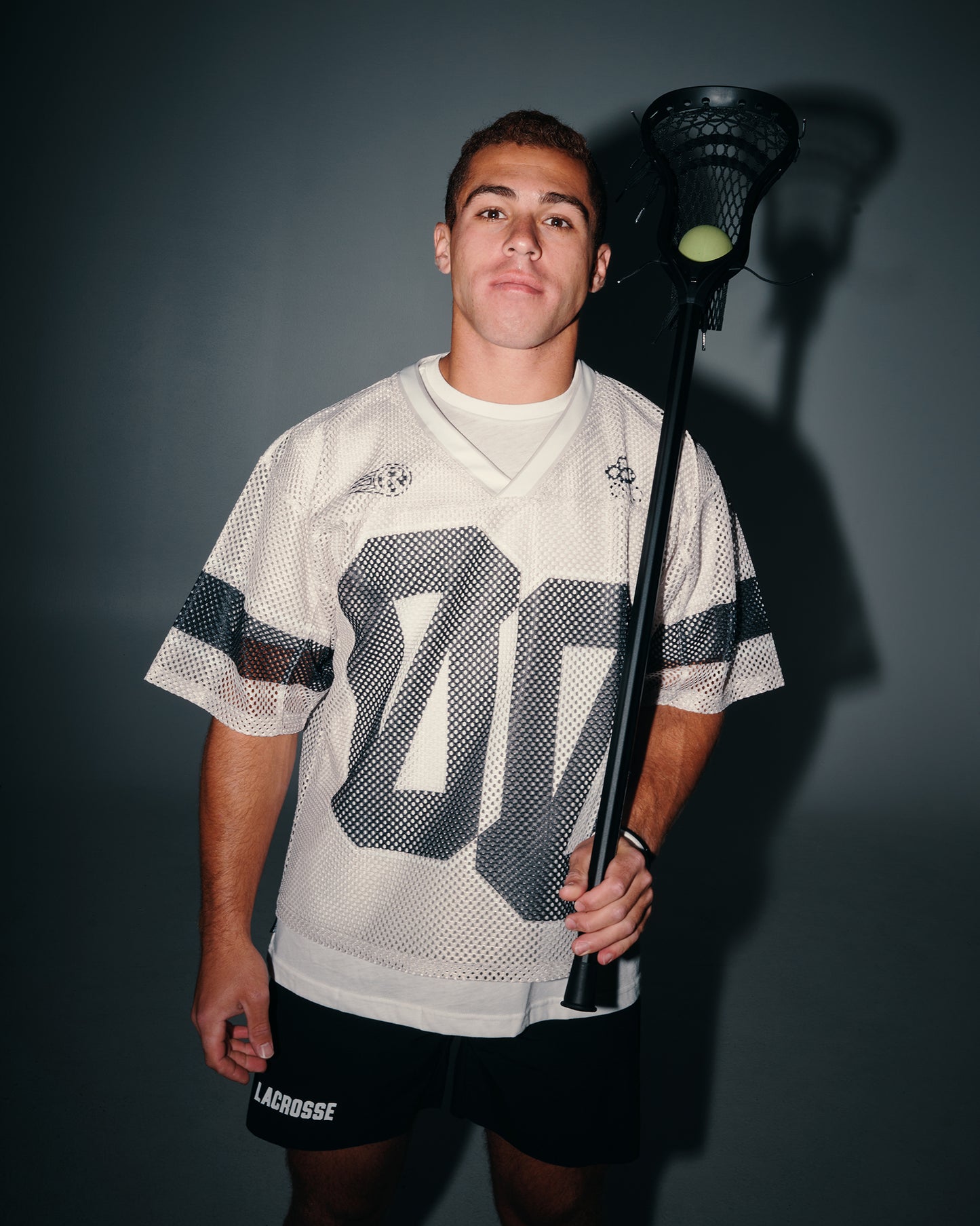 A young man wearing a white lacrosse jersey with a large gray number 00 on the front, black lacrosse shorts, and holding a lacrosse stick with a ball, posed against a dark gray background.