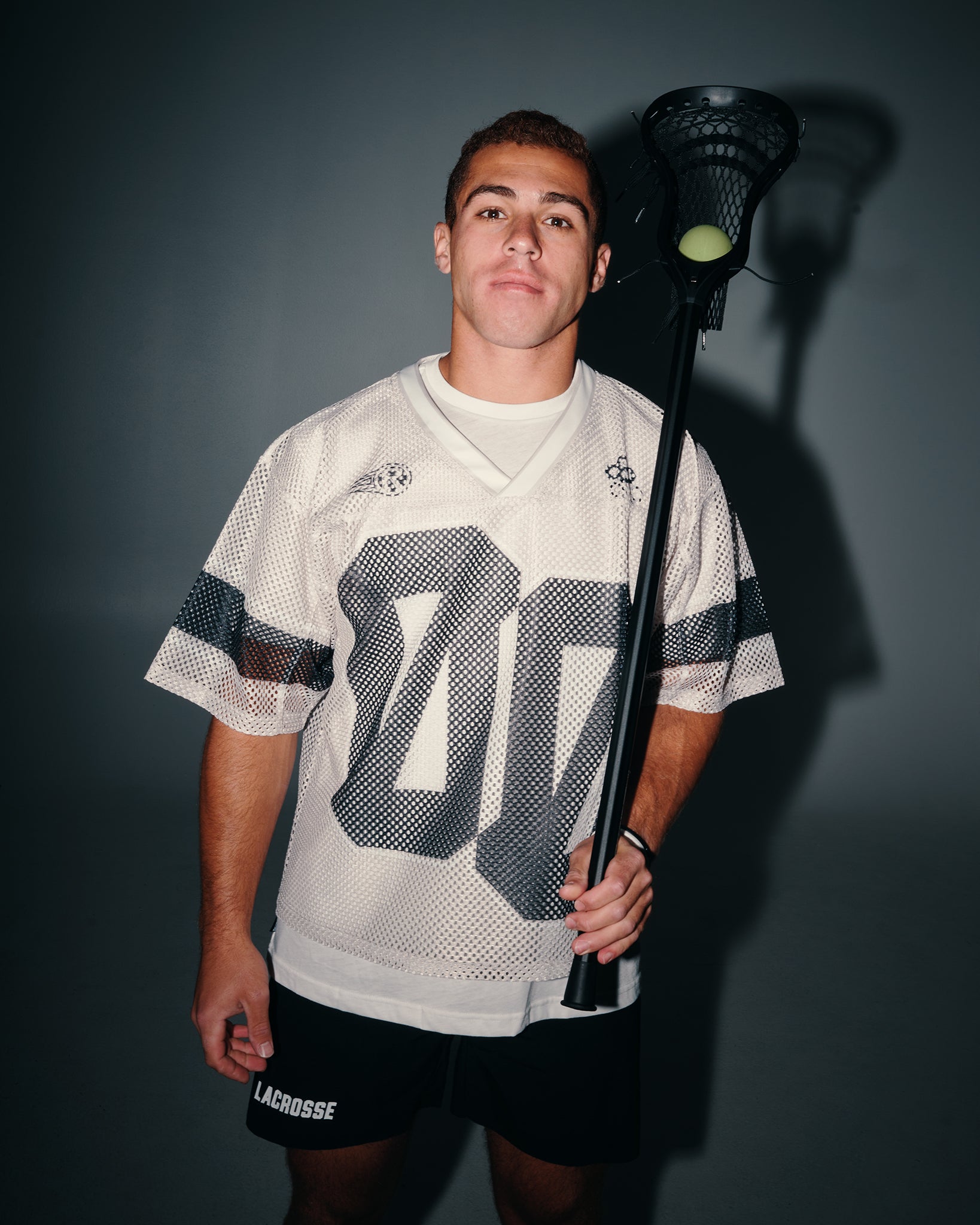 A young man wearing a white lacrosse jersey with a large gray number 00 on the front, black lacrosse shorts, and holding a lacrosse stick with a ball, posed against a dark gray background.