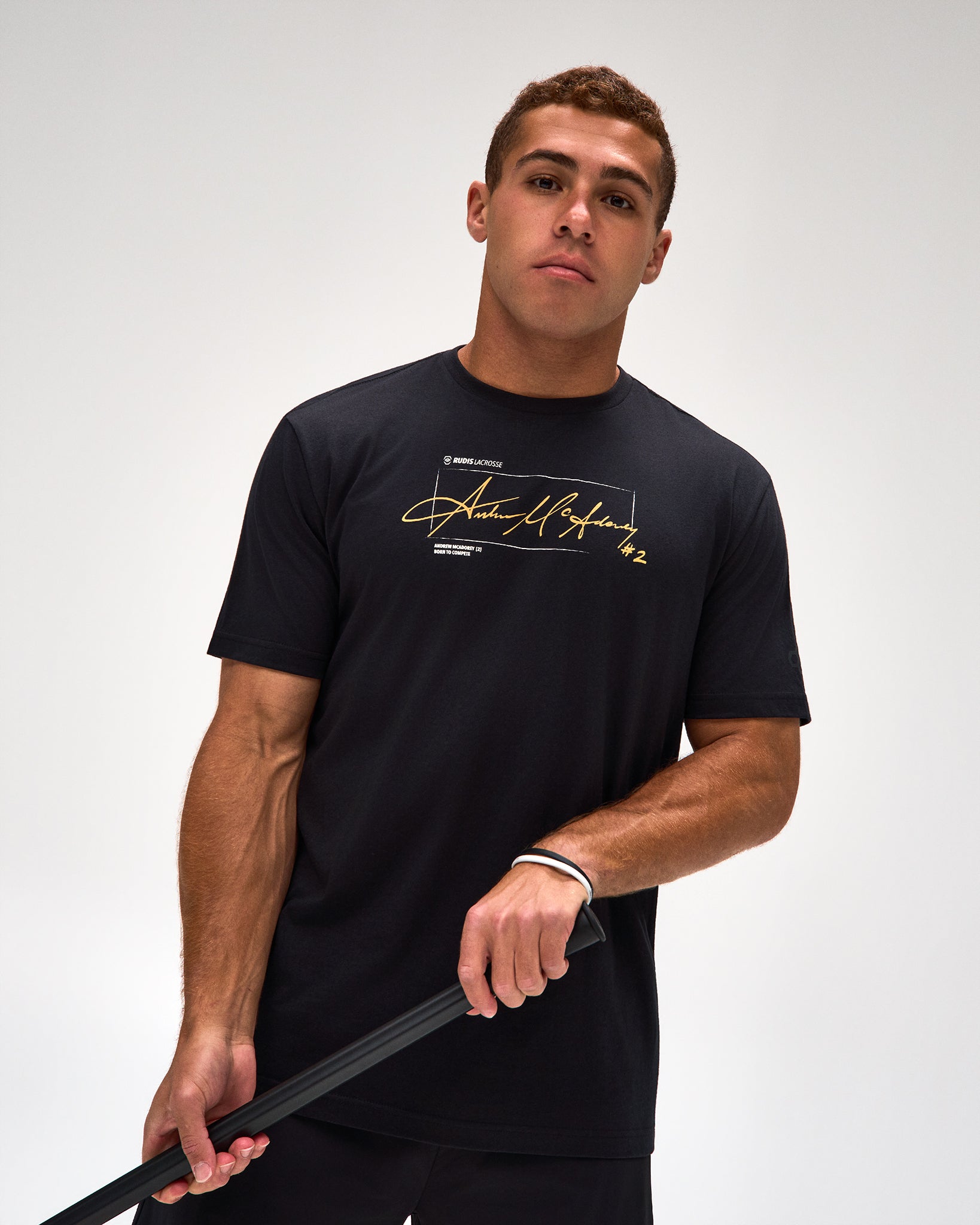A man, Andrew McAdorey, wearing a black short-sleeve t-shirt with a gold signature graphic on the chest, holding a lacrosse stick against a light gray studio background.