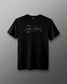 A black short-sleeve crewneck t-shirt featuring a gold cursive signature graphic and small white text on the chest, displayed against a dark gray gradient background.