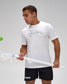 A man wearing a white Lacoste t-shirt with a green signature graphic and black lacrosse shorts, holding a lacrosse stick while looking at a green ball against a light gray background.