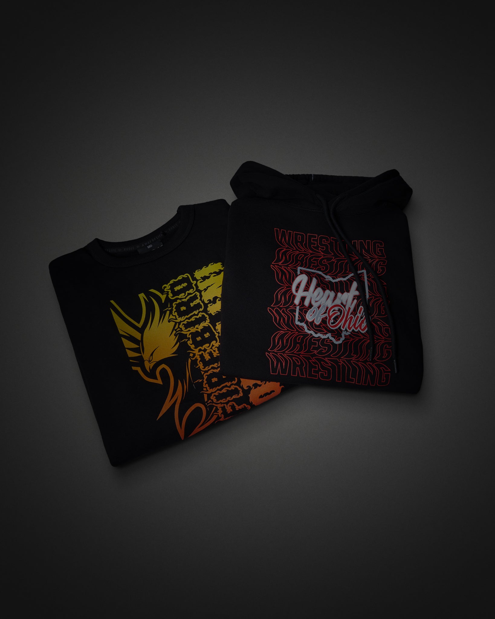 Two folded black Hoodie and Crewneck displayed on a dark gray gradient background. The left shirt features a yellow, orange, and red graphic of a winged skeletal figure.