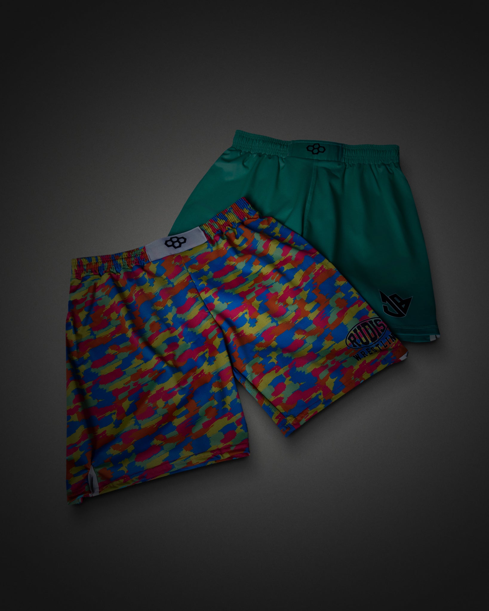 Two pairs of athletic shorts displayed together on a dark gray background. The front pair features a vibrant multicolor camouflage pattern in red, blue, yellow, and green, while the back pair is a solid teal green with a small black logo on the side.