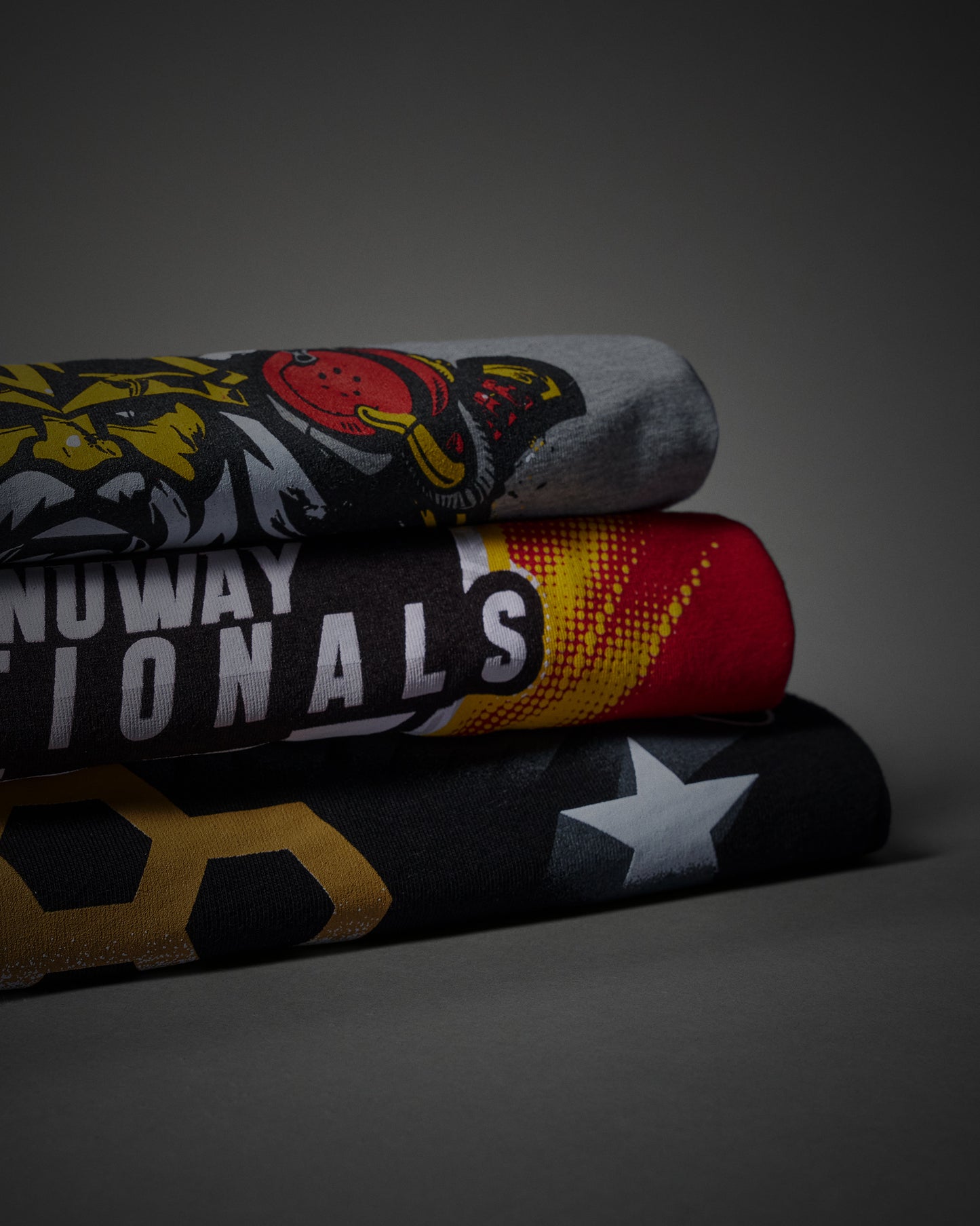 Three folded athletic t-shirts stacked on a dark gray background. The top t-shirt is heather gray with a yellow and red tiger graphic.