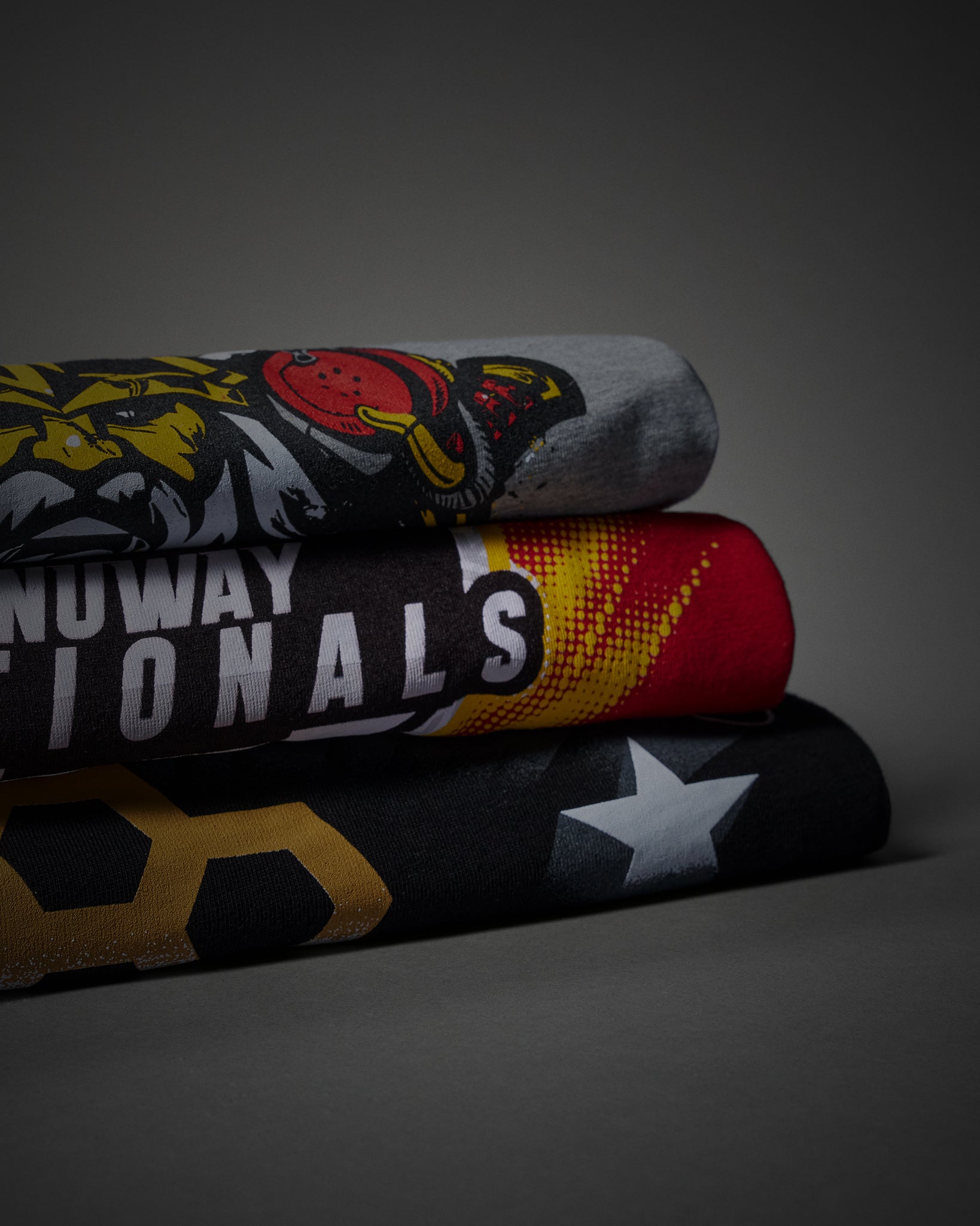 Three folded athletic t-shirts stacked on a dark gray background. The top t-shirt is heather gray with a yellow and red tiger graphic.