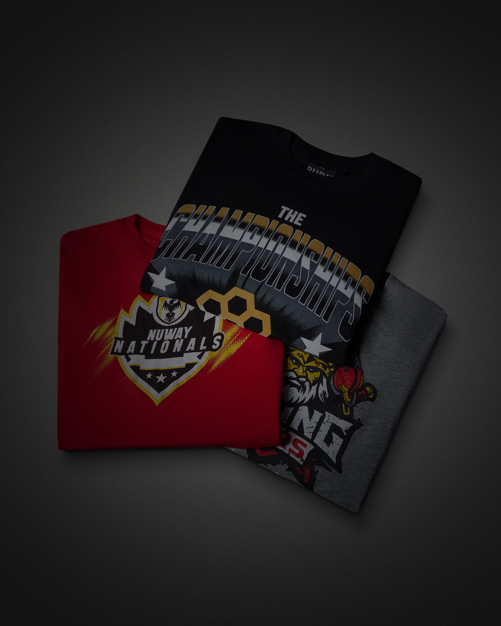 Three folded graphic T-shirts arranged in an overlapping stack on a dark gray background. The top shirt is black with a large white, gray, and gold championship graphic reading 'The Championship'; the middle shirt is red with a large 'Hawkey Nationals' logo in gold, black, and white; the bottom shirt is dark gray with a tiger graphic and partial text.