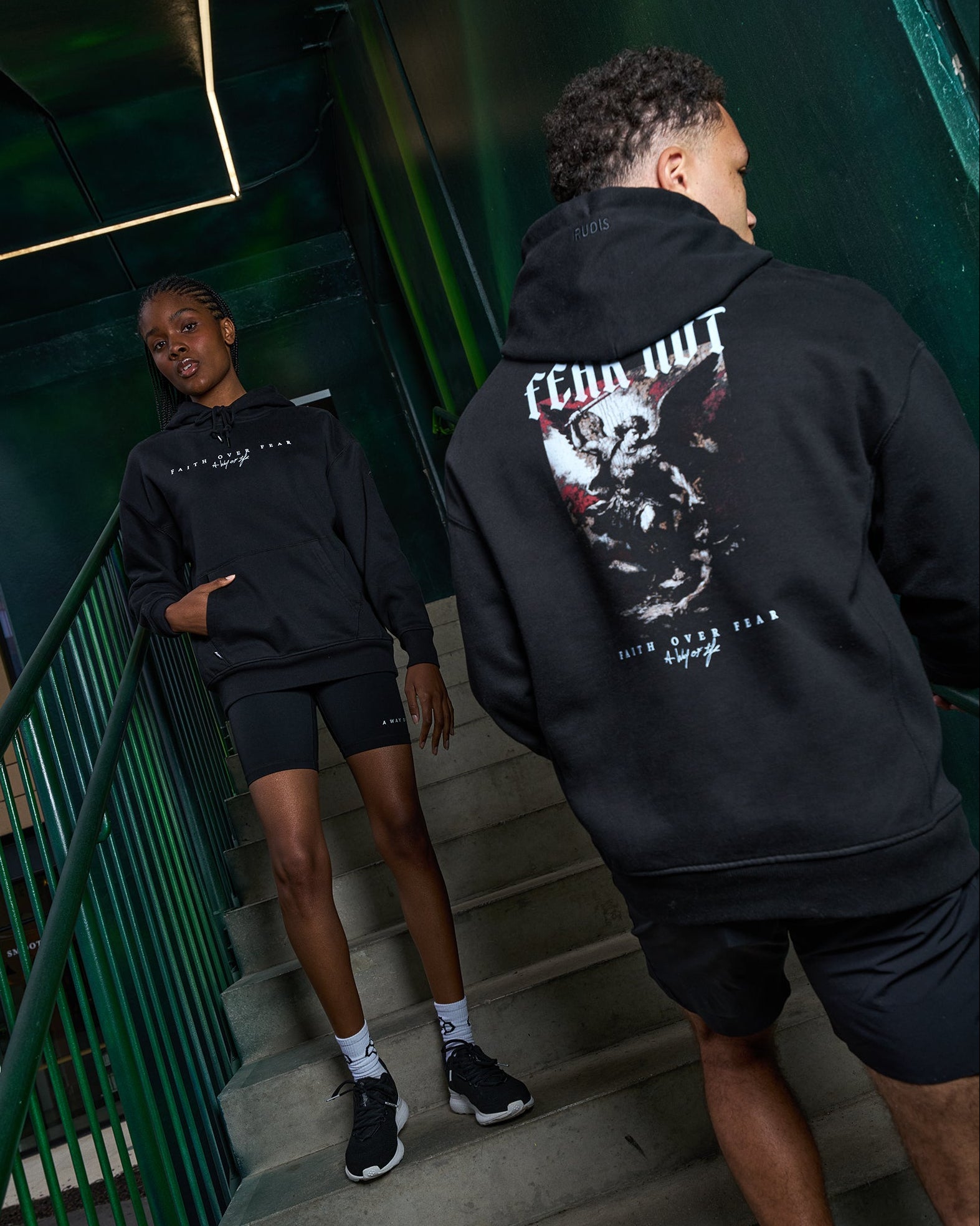 Two people wearing black RUDIS hoodies with graphic designs on a staircase.