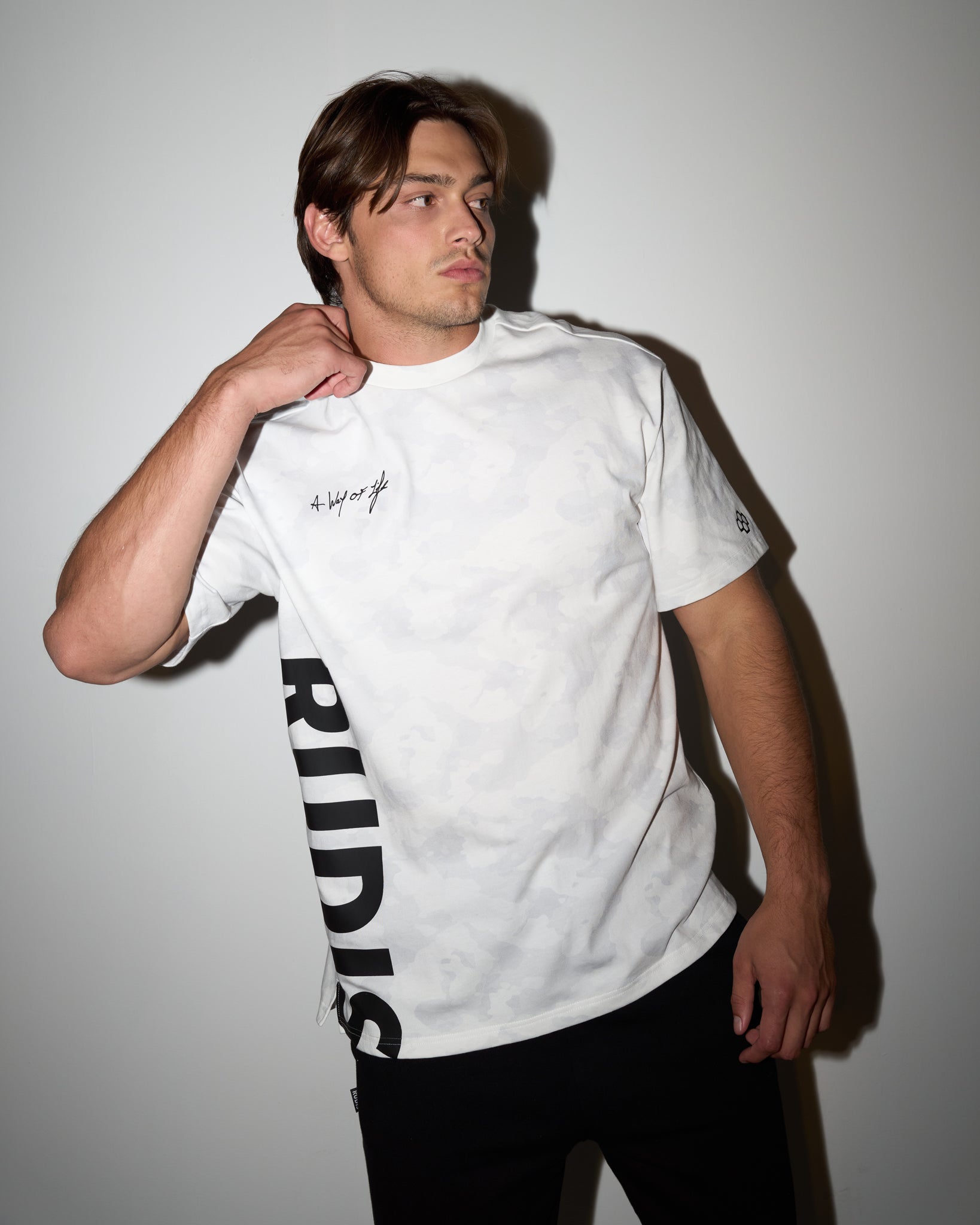 Model wearing the RUDIS A Way of Life Tee in white camo, featuring bold vertical RUDIS lettering along the side and script detail on the chest—styled in a modern athletic fit under direct light.