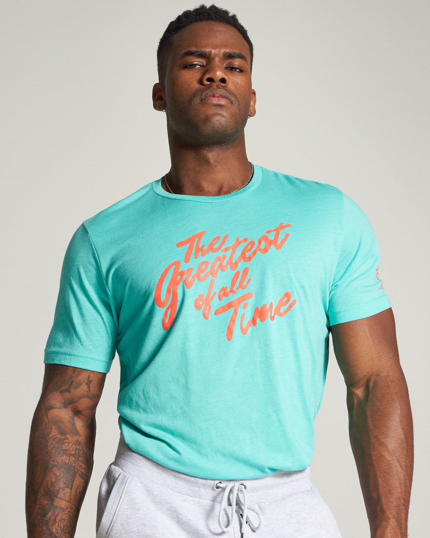 A muscular man poses confidently in a turquoise t-shirt featuring the slogan The Greatest of All Time in bold orange text complemented by gray sweatpants