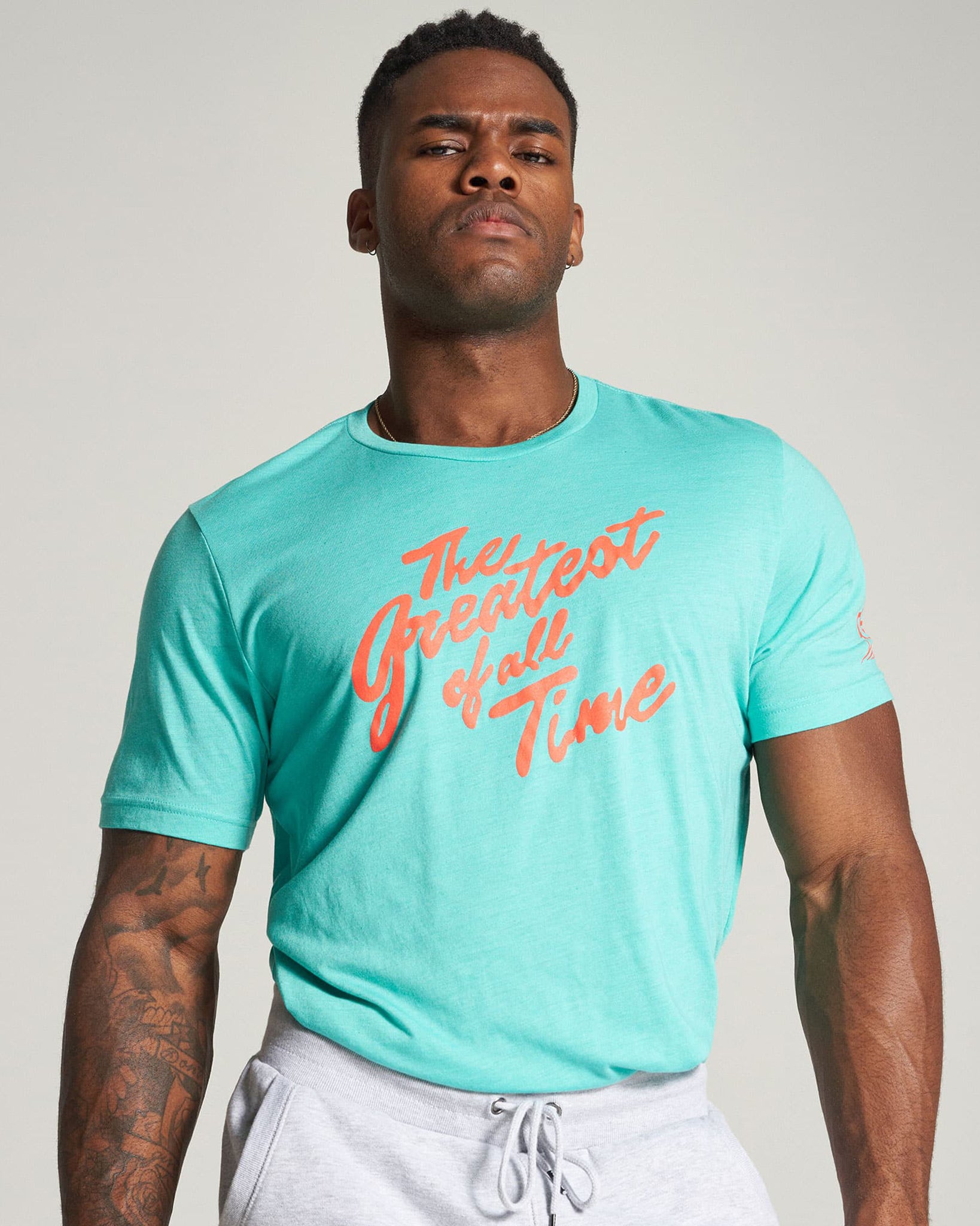 A muscular man poses confidently in a turquoise t-shirt featuring the slogan The Greatest of All Time in bold orange text complemented by gray sweatpants