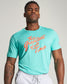 A muscular man poses confidently in a turquoise t-shirt featuring the slogan The Greatest of All Time in bold orange text complemented by gray sweatpants