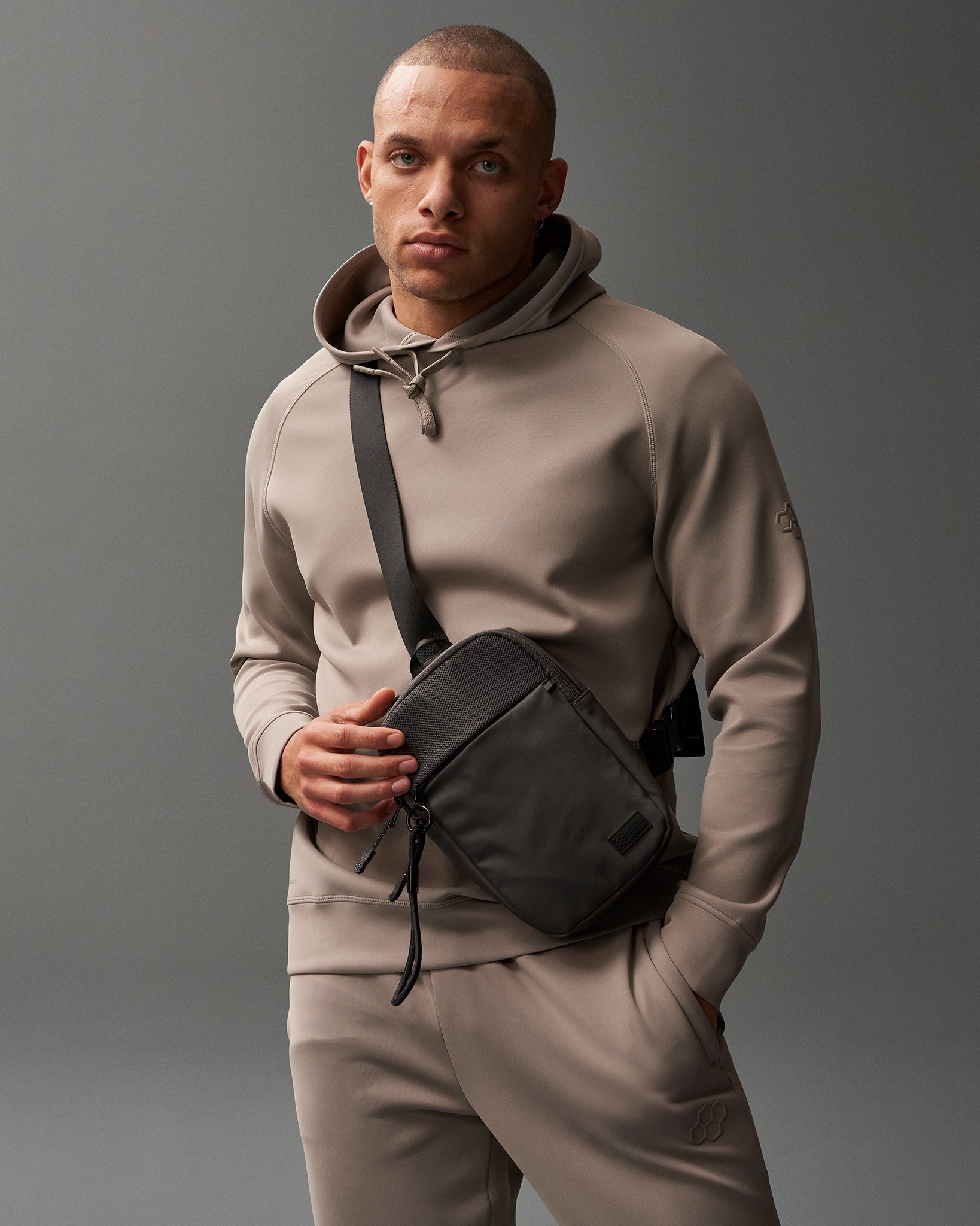 A male figure in a beige hoodie and joggers stands against a gray background, holding a black crossbody bag with a textured flap, presenting a stylish and casual appearance.