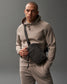 A male figure in a beige hoodie and joggers stands against a gray background, holding a black crossbody bag with a textured flap, presenting a stylish and casual appearance.