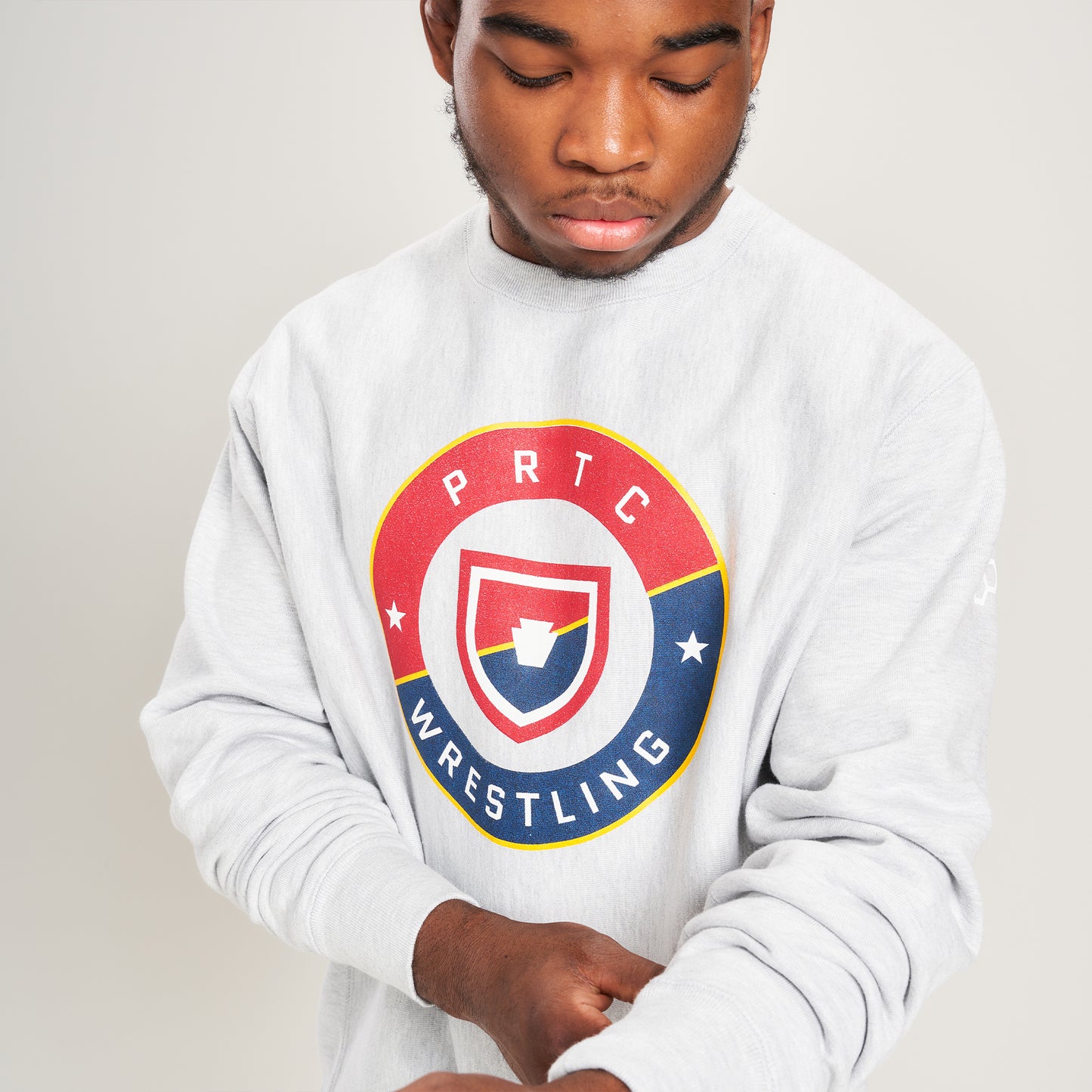 A young man in a gray sweatshirt with a bold PRTC Wrestling logo adjusts the cuff displaying a focused expression against a simple background