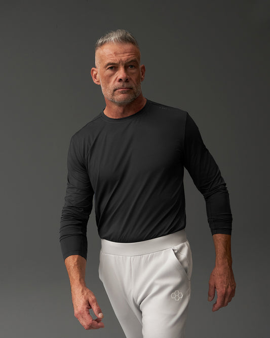 A middle-aged man in a black long-sleeve shirt and light gray pants stands against a dark gray background, showcasing a stylish and athletic look.