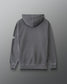 A dark gray pullover hoodie shown from the back, featuring a small white geometric logo on the left sleeve, set against a dark gray gradient background.