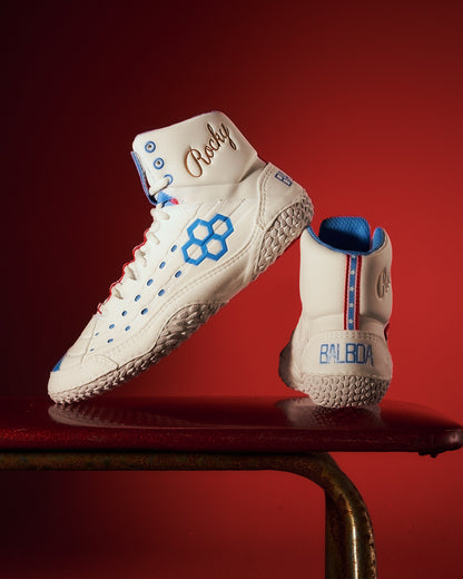 Classic white high-top boxing shoes featuring blue accents and the name Rocky embossed on the ankle displayed against a striking red background