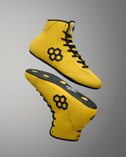 Yellow wrestling shoes with black accents on a gray background. RUDIS Hex Logo Rocky Shoes