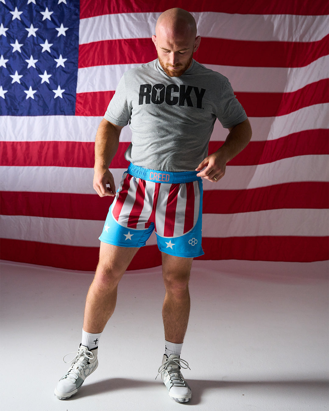 Man wearing a 'Rocky' t-shirt and American flag-themed shorts in front of an American flag.