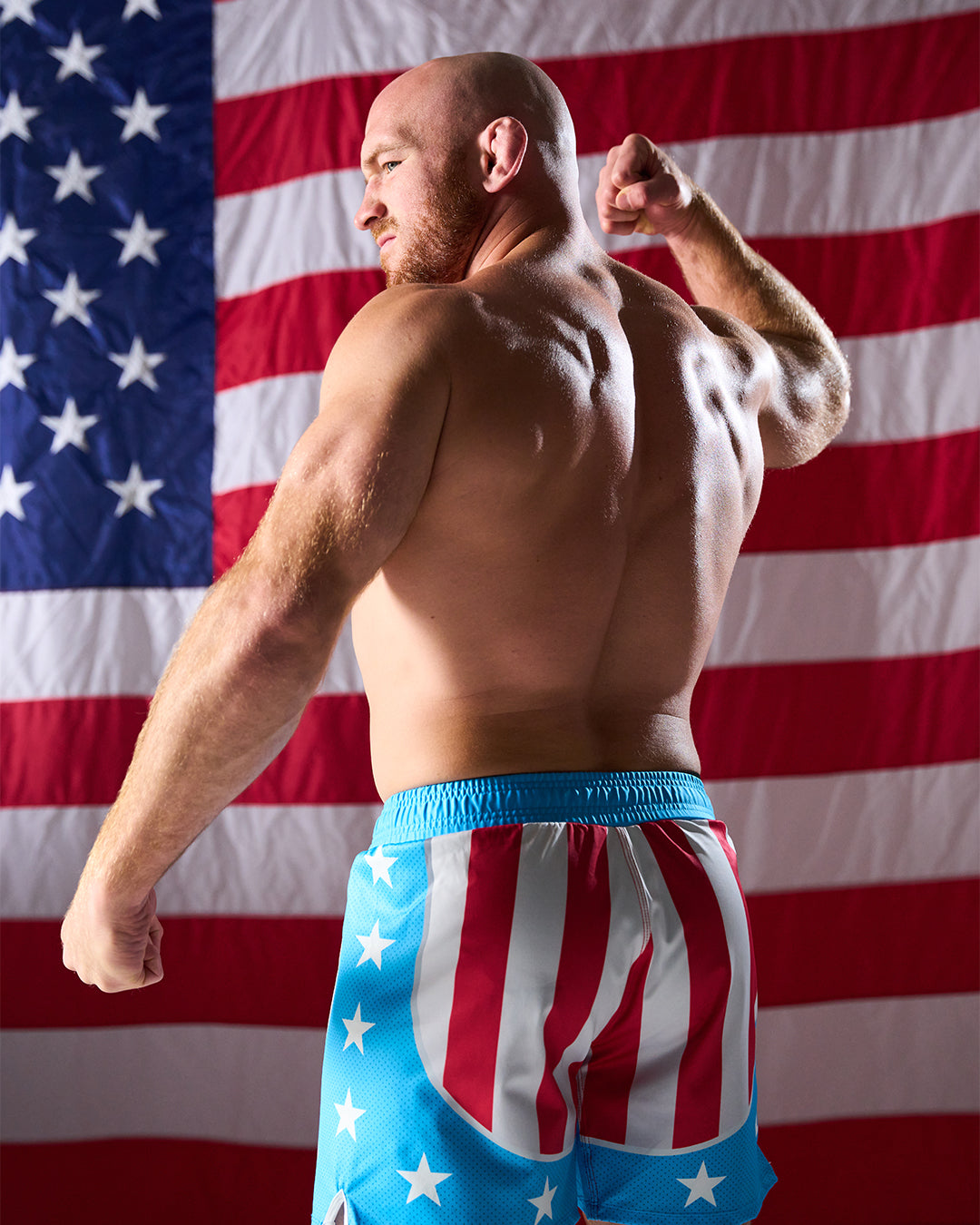 Man flexing muscles wearing American flag-themed shorts with a U.S. flag in the background