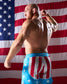 Man flexing muscles wearing American flag-themed shorts with a U.S. flag in the background