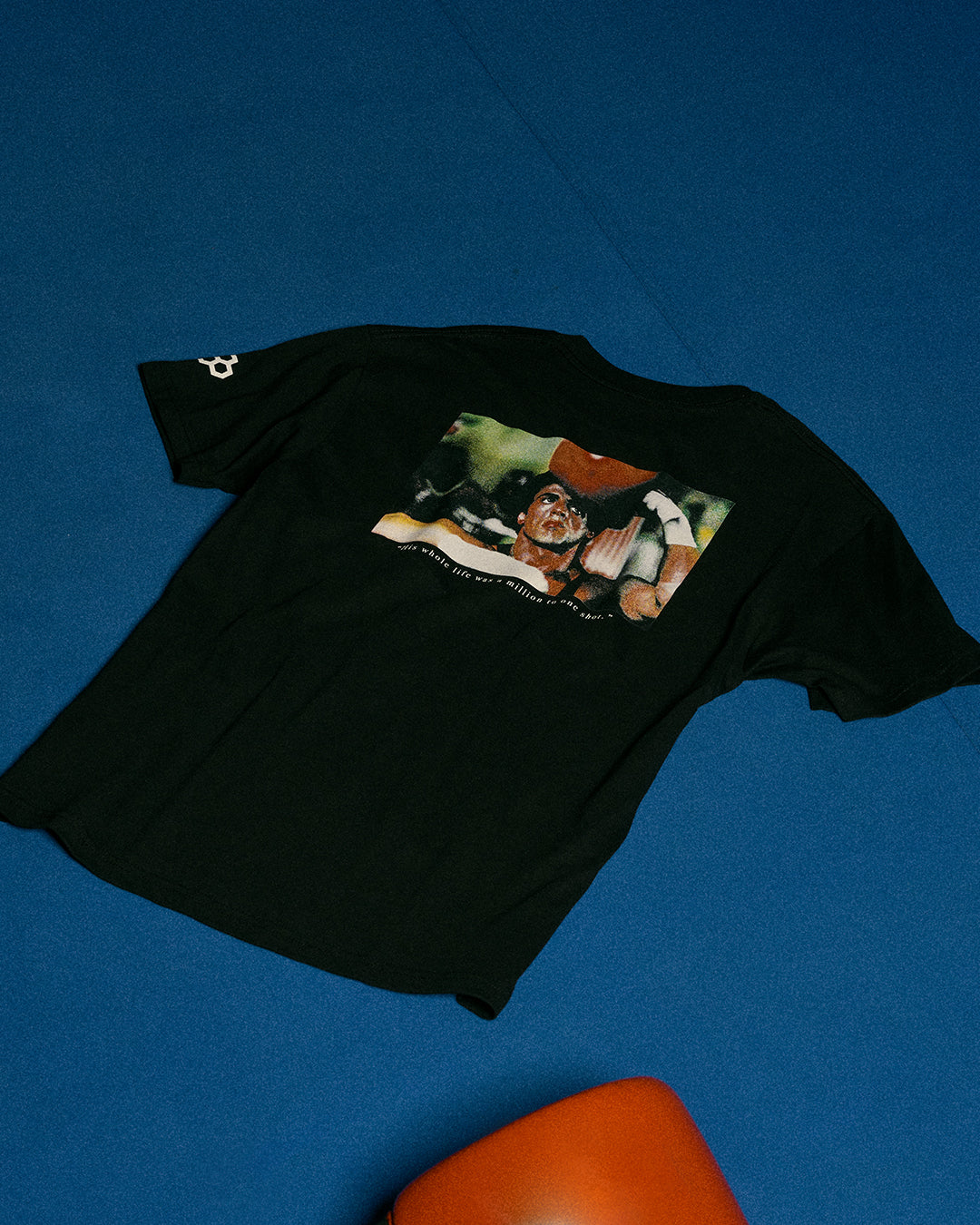 A black short-sleeve graphic t-shirt laid flat on a textured blue background, featuring a large multicolor print on the back depicting a boxing scene.