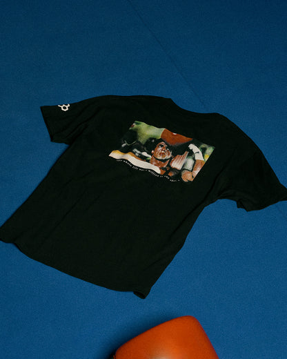 A black short-sleeve graphic t-shirt laid flat on a textured blue background, featuring a large multicolor print on the back depicting a boxing scene.