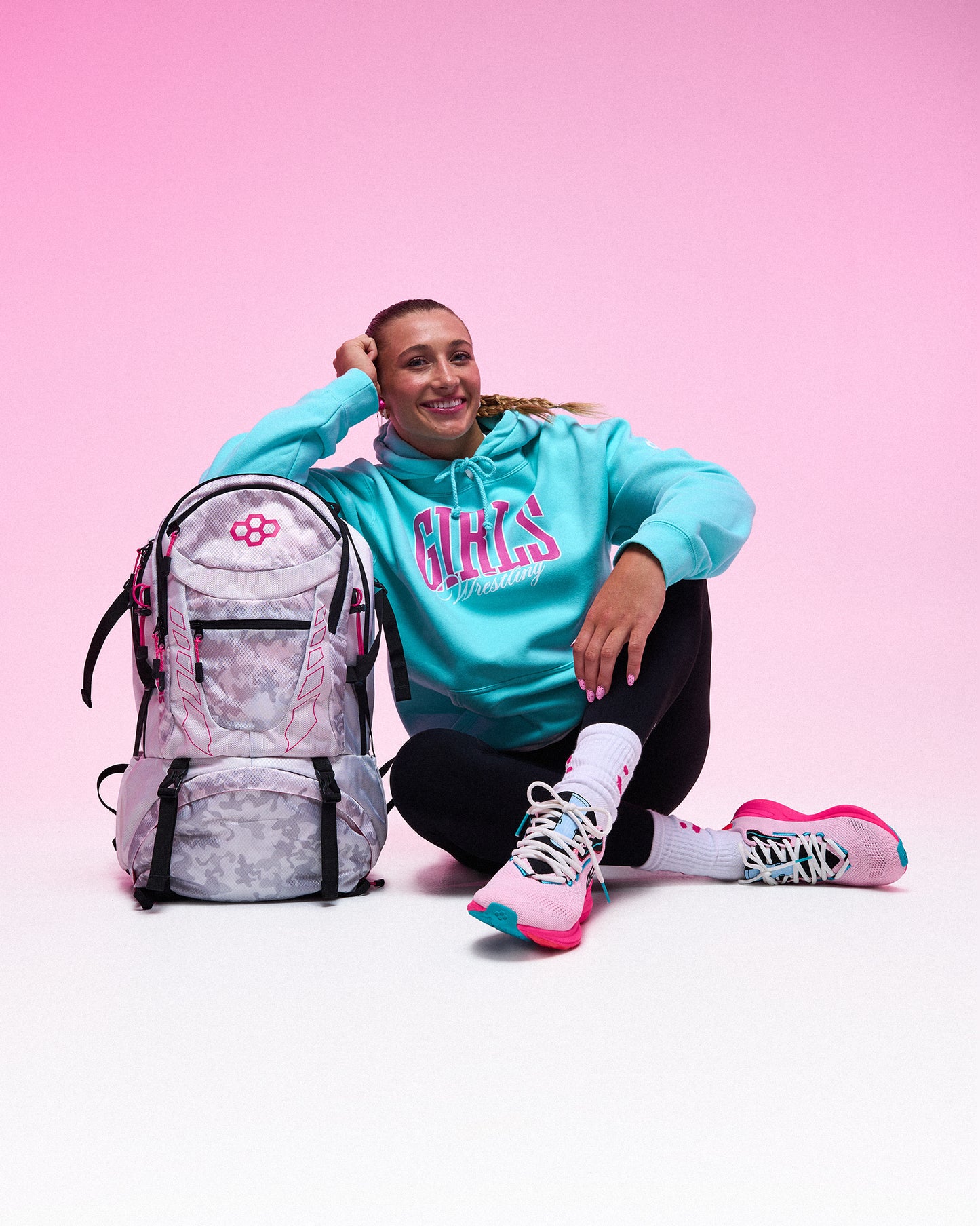 A woman sitting on a white studio floor next to a pink and gray patterned backpack, wearing a turquoise hoodie with pink 'GIRLS' lettering, black leggings, and pink sneakers against a pink gradient background.