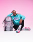 A woman sitting on a white studio floor next to a pink and gray patterned backpack, wearing a turquoise hoodie with pink 'GIRLS' lettering, black leggings, and pink sneakers against a pink gradient background.