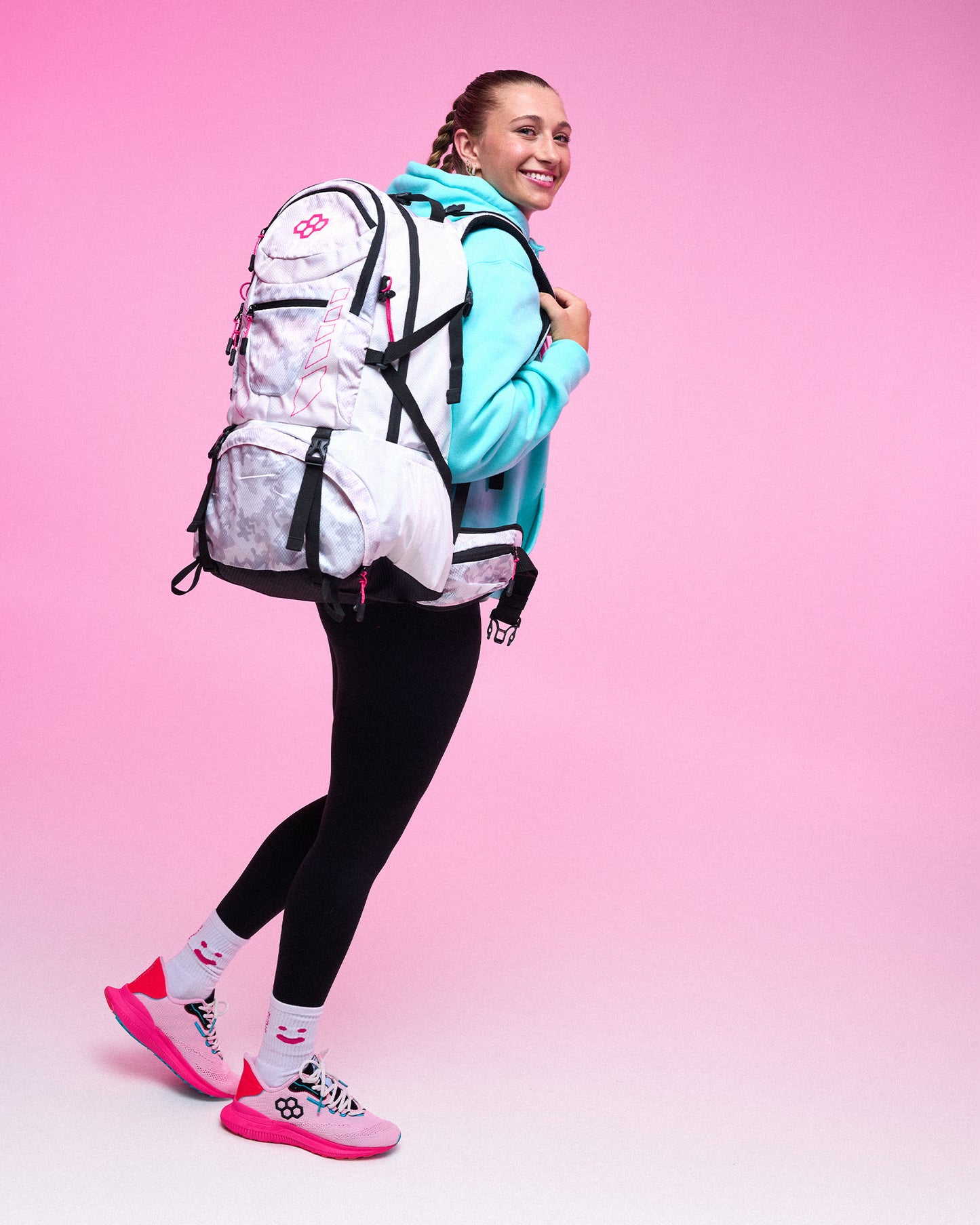 A woman posing with a large white and pink backpack, wearing a light blue hoodie, black leggings, and pink and white sneakers, set against a solid pink studio background.