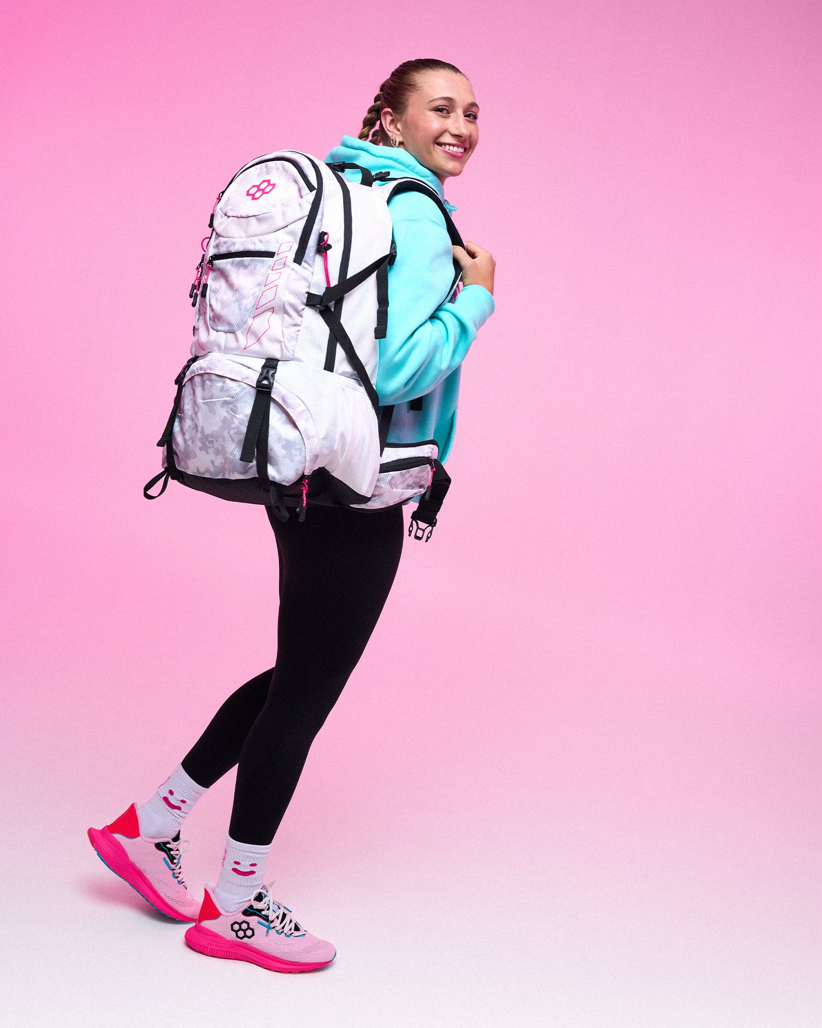A woman posing with a large white and pink backpack, wearing a light blue hoodie, black leggings, and pink and white sneakers, set against a solid pink studio background.