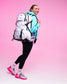 A woman posing with a large white and pink backpack, wearing a light blue hoodie, black leggings, and pink and white sneakers, set against a solid pink studio background.