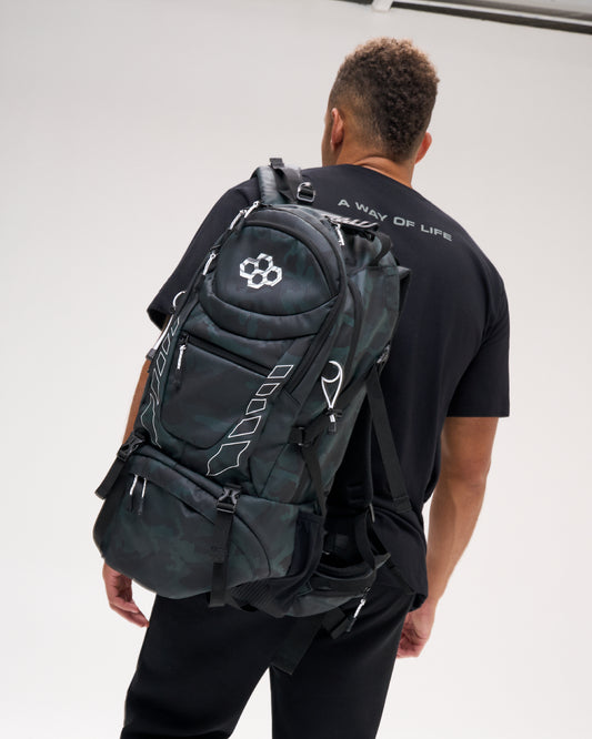 RUDIS Genesis Gearpack IV in Green Camo worn over one shoulder, showcasing the RUDIS logo, multiple zippered compartments, compression straps, side storage, and structured wrestling gear storage design.