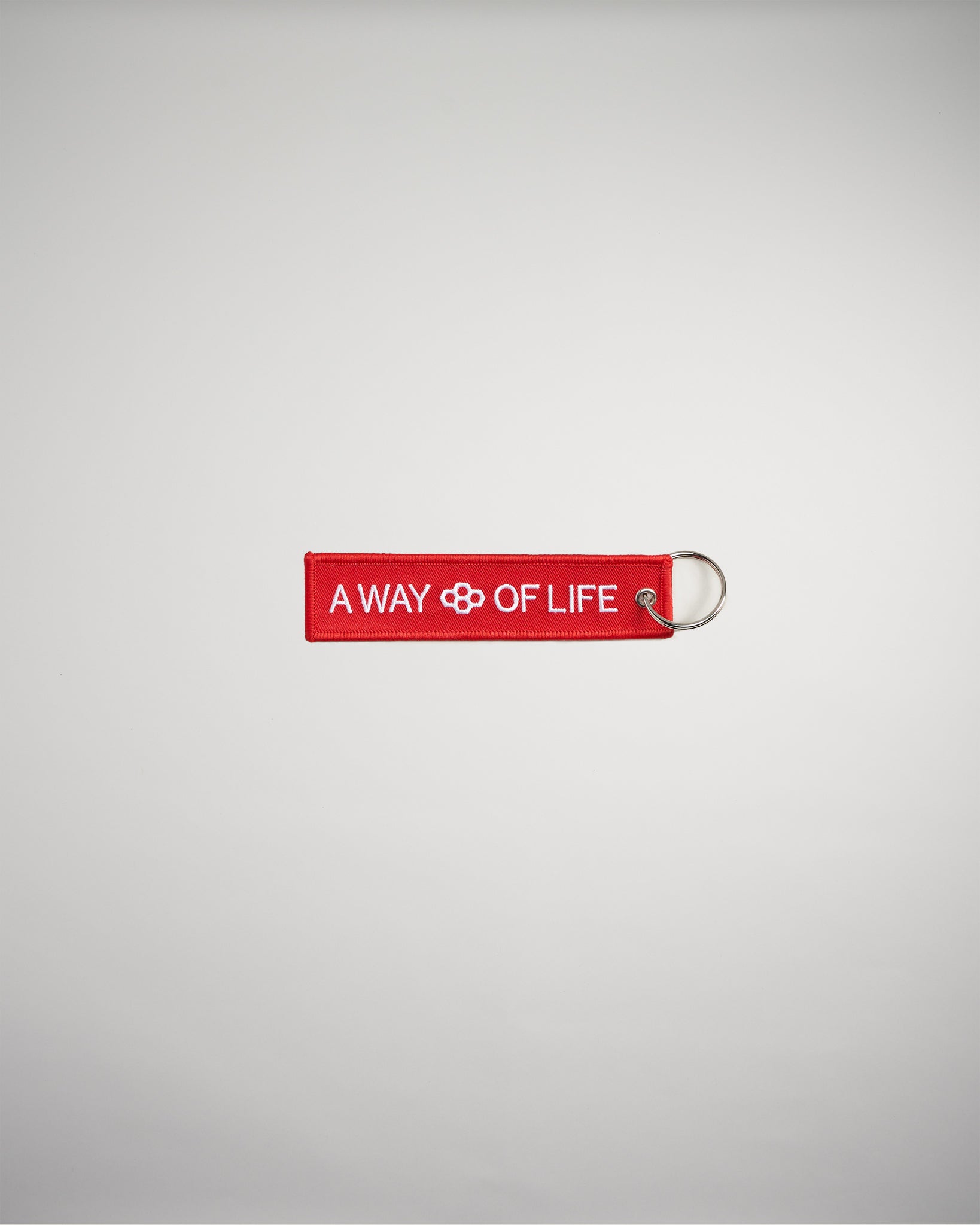 A red keychain tag featuring the phrase A WAY OF LIFE with a decorative symbol in the center designed to convey a lifestyle message
