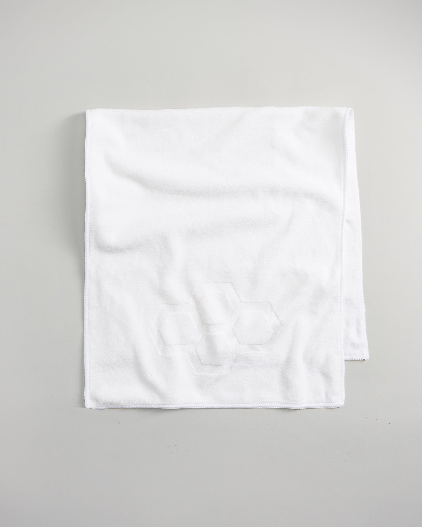 A soft white rectangular towel featuring a subtle hexagonal pattern in the corner resting on a neutral background