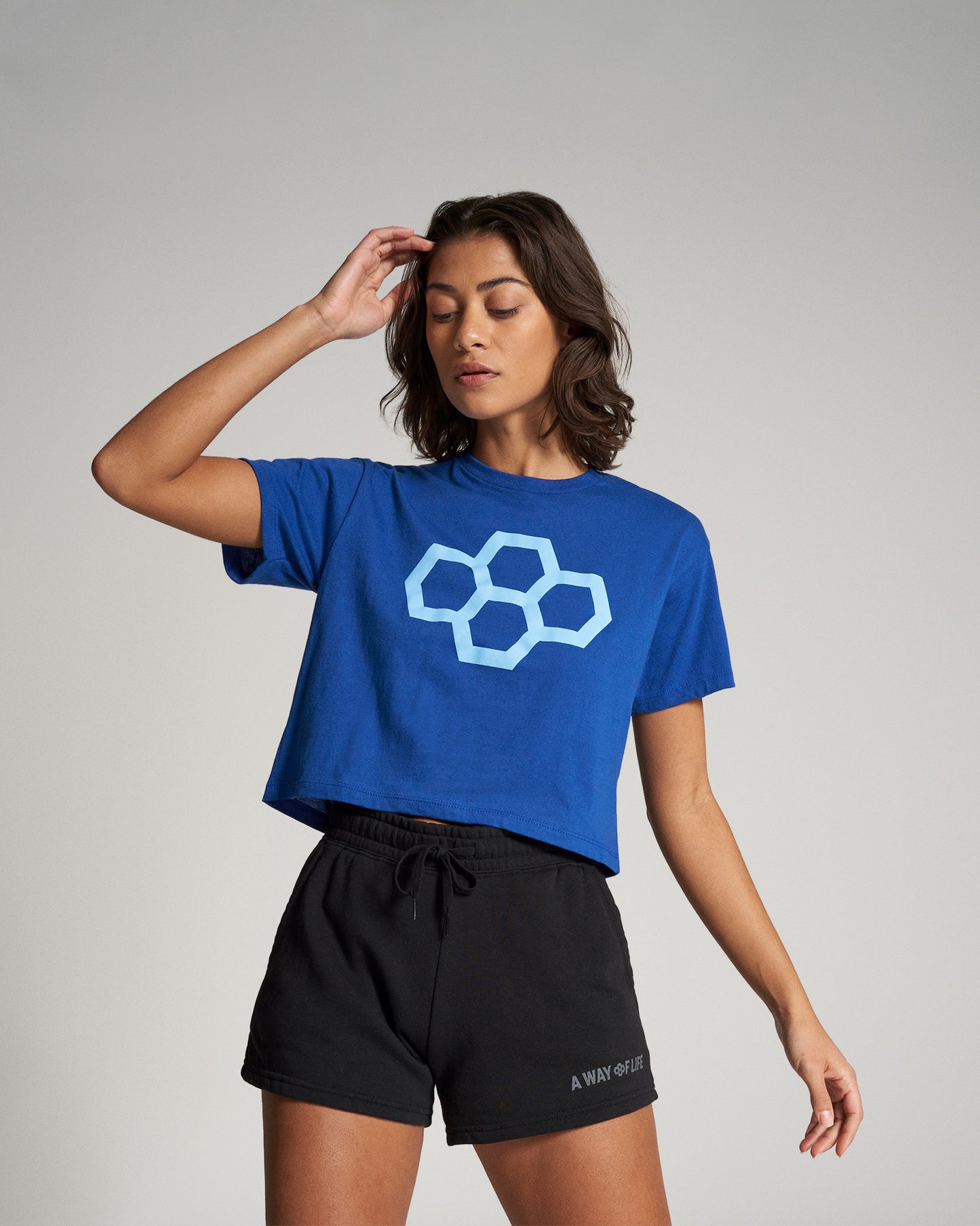 A young woman is modeling a blue cropped t-shirt with a geometric logo and black shorts set against a neutral background