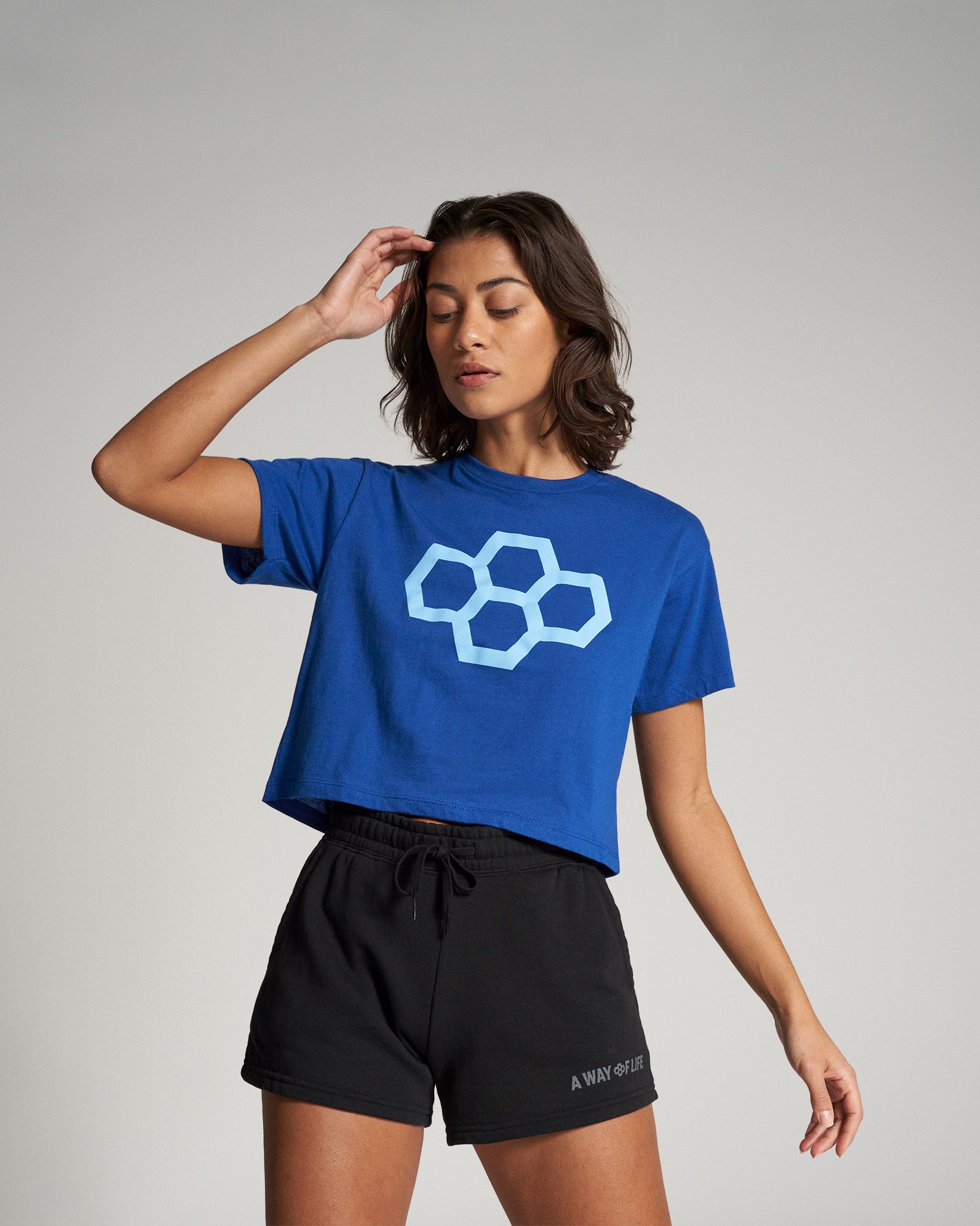 A young woman is modeling a blue cropped t-shirt with a geometric logo and black shorts set against a neutral background