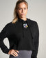 A woman poses confidently in a cropped black hoodie featuring a colorful design on the chest paired with gray leggings and a neutral background