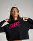A close-up portrait of a woman in a stylish black cropped hoodie featuring bright pink lettering that reads RUDIS set against a neutral background