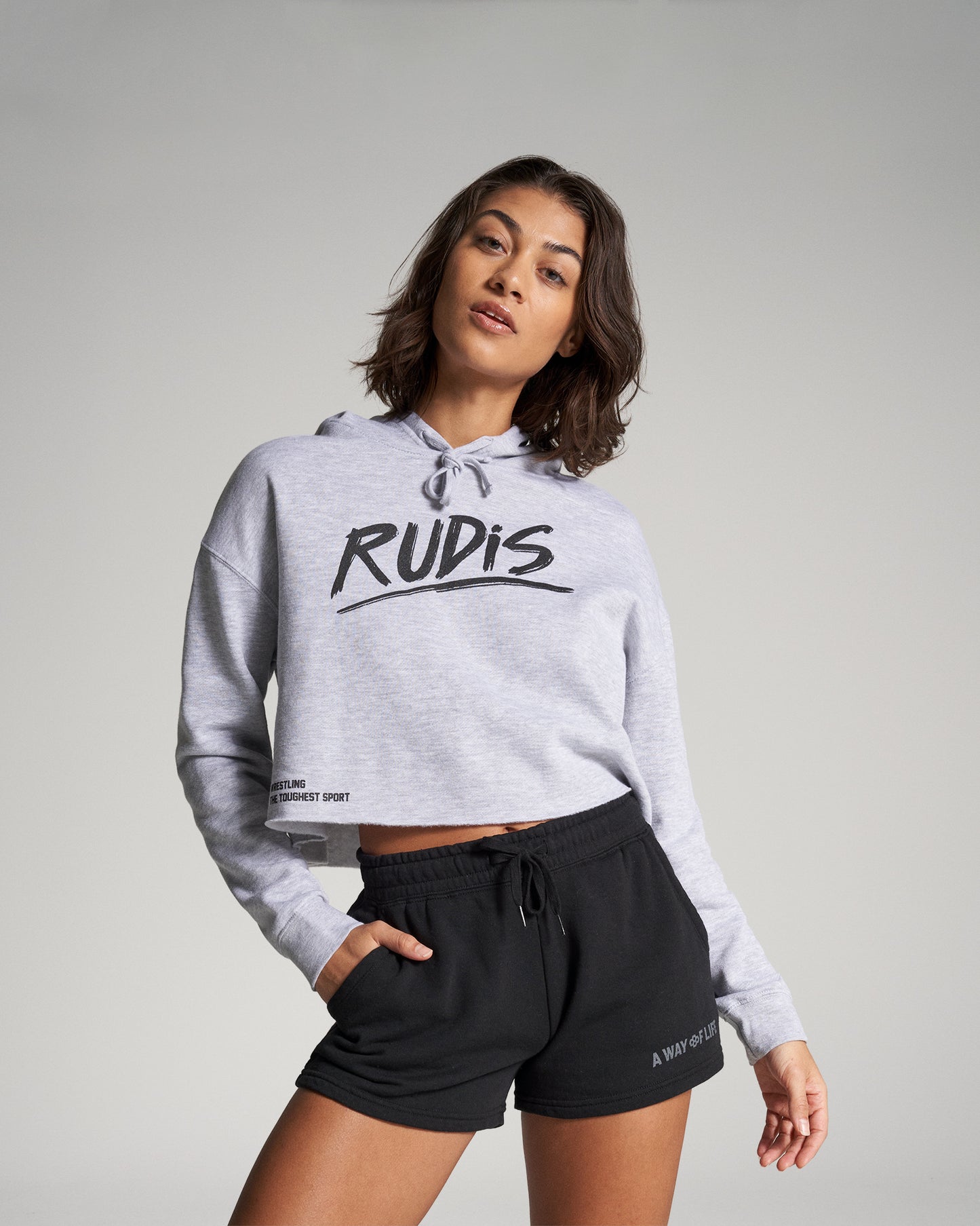 A young woman in a grey cropped hoodie with RUDIS printed on it and black shorts poses confidently with her hands on her hips against a neutral background