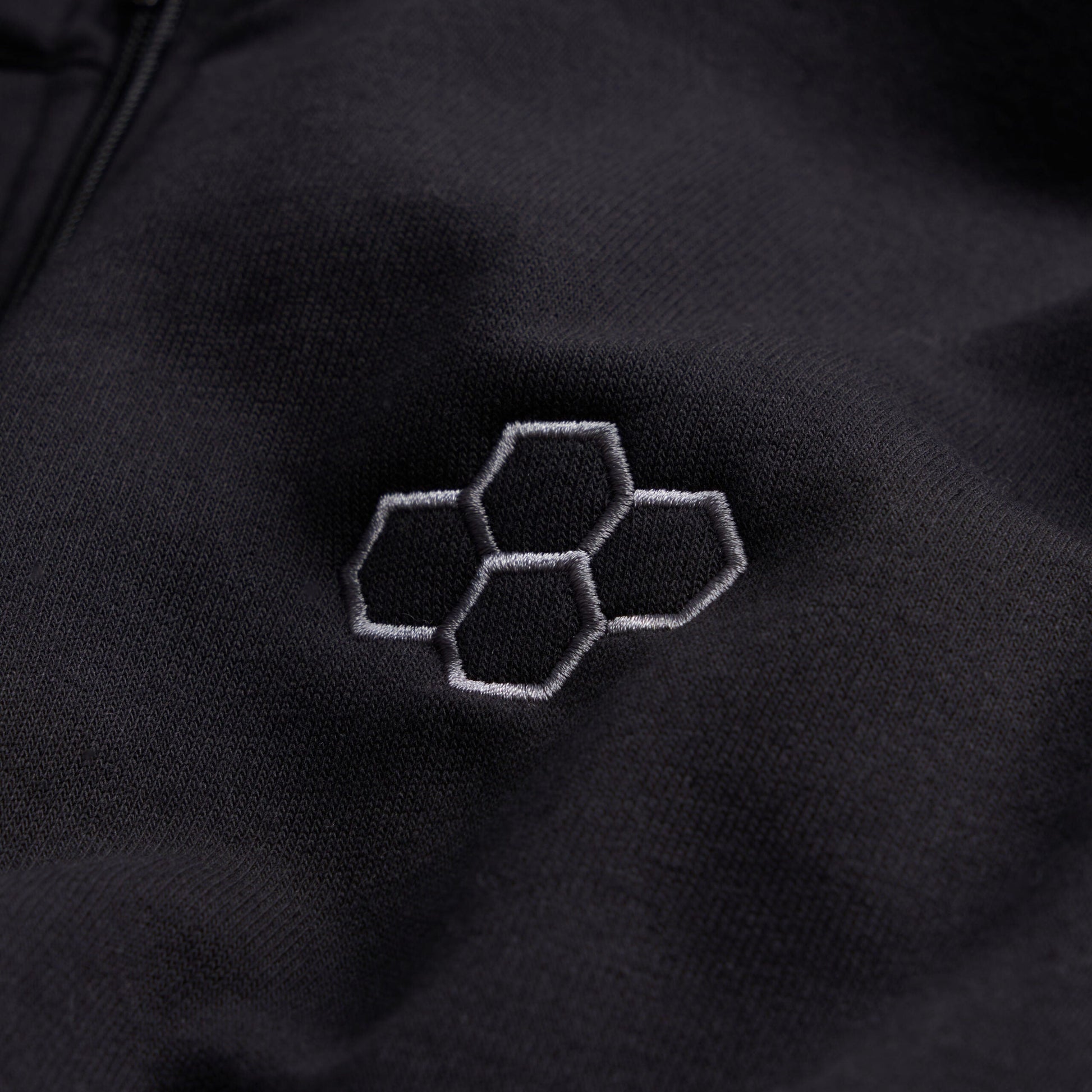 This image features a close-up of a black fabric surface adorned with an embroidered hexagonal logo in a lighter gray tone showcasing a sleek and modern design aesthetic