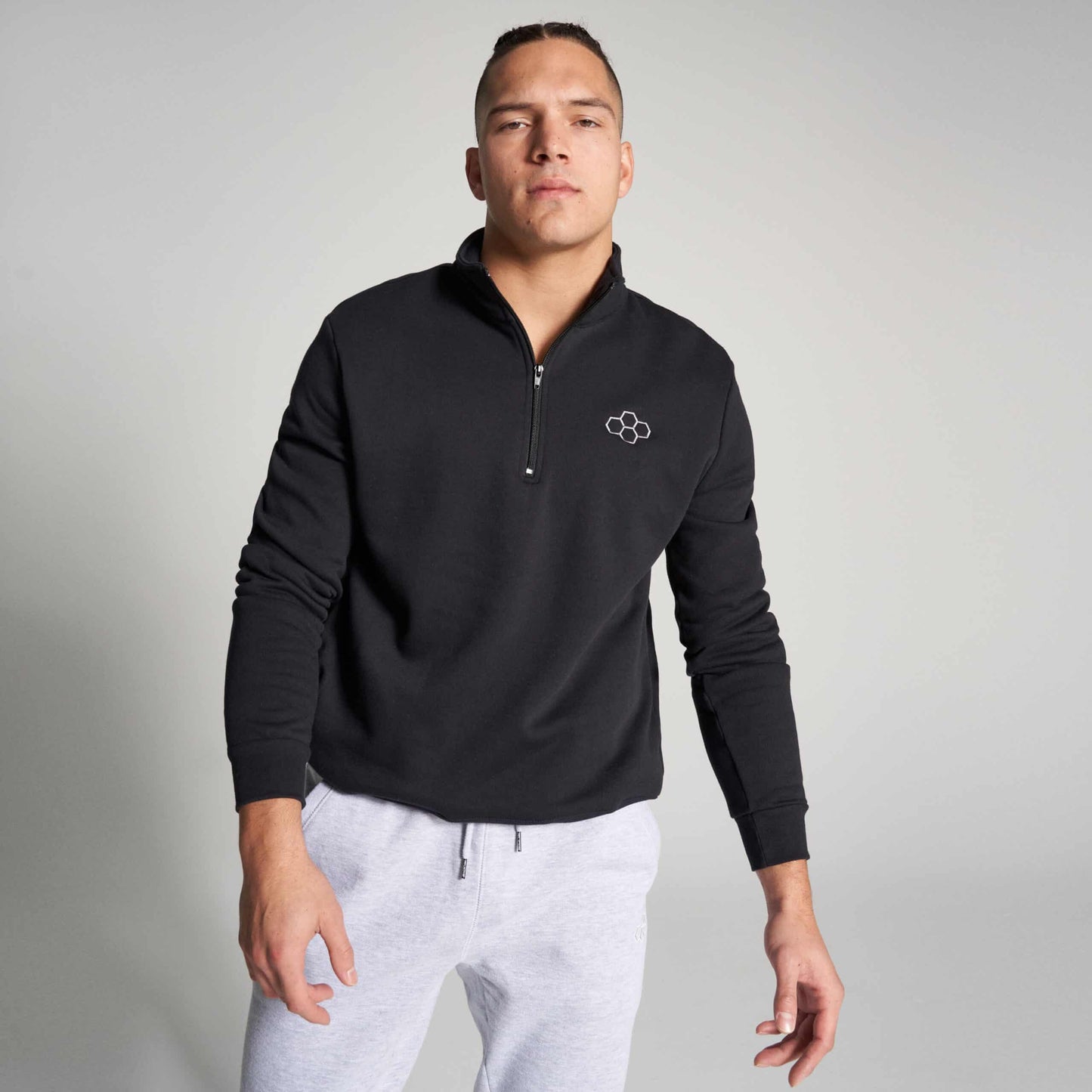 A confident male model wears a black zip-up sweatshirt with a minimalist logo paired with light gray sweatpants set against a neutral background