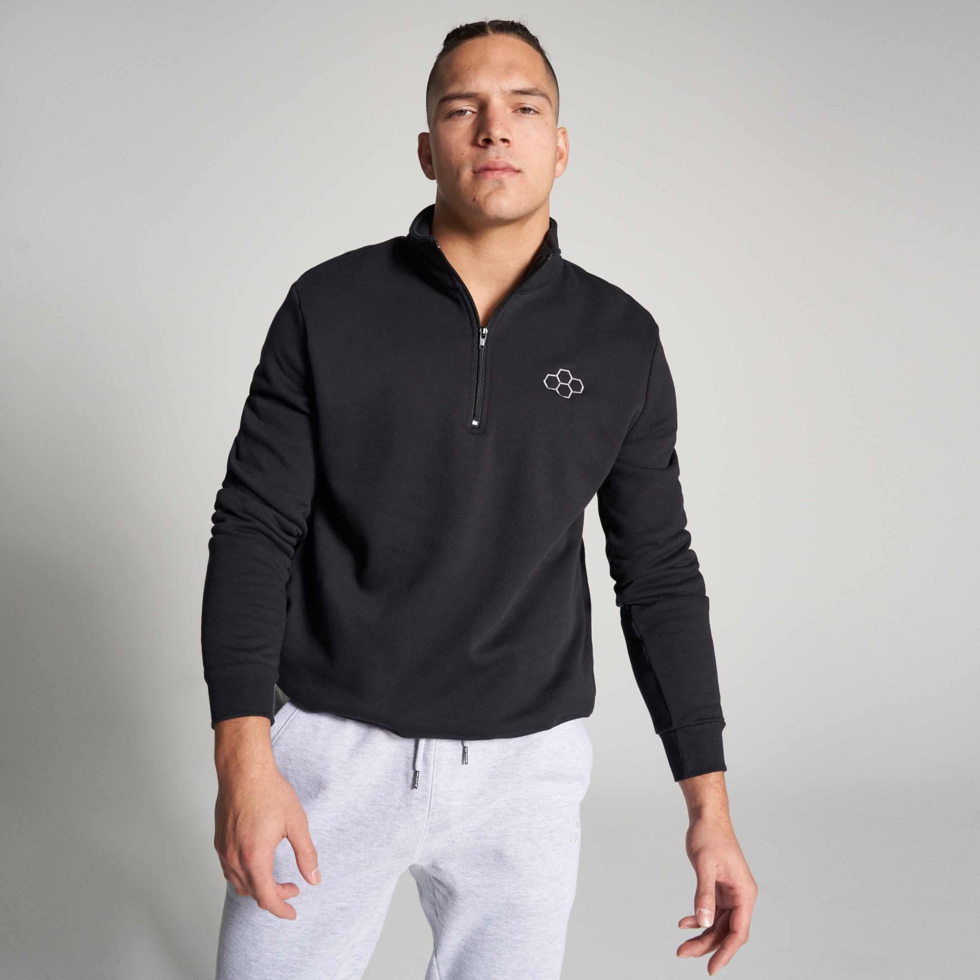 A confident male model wears a black zip-up sweatshirt with a minimalist logo paired with light gray sweatpants set against a neutral background