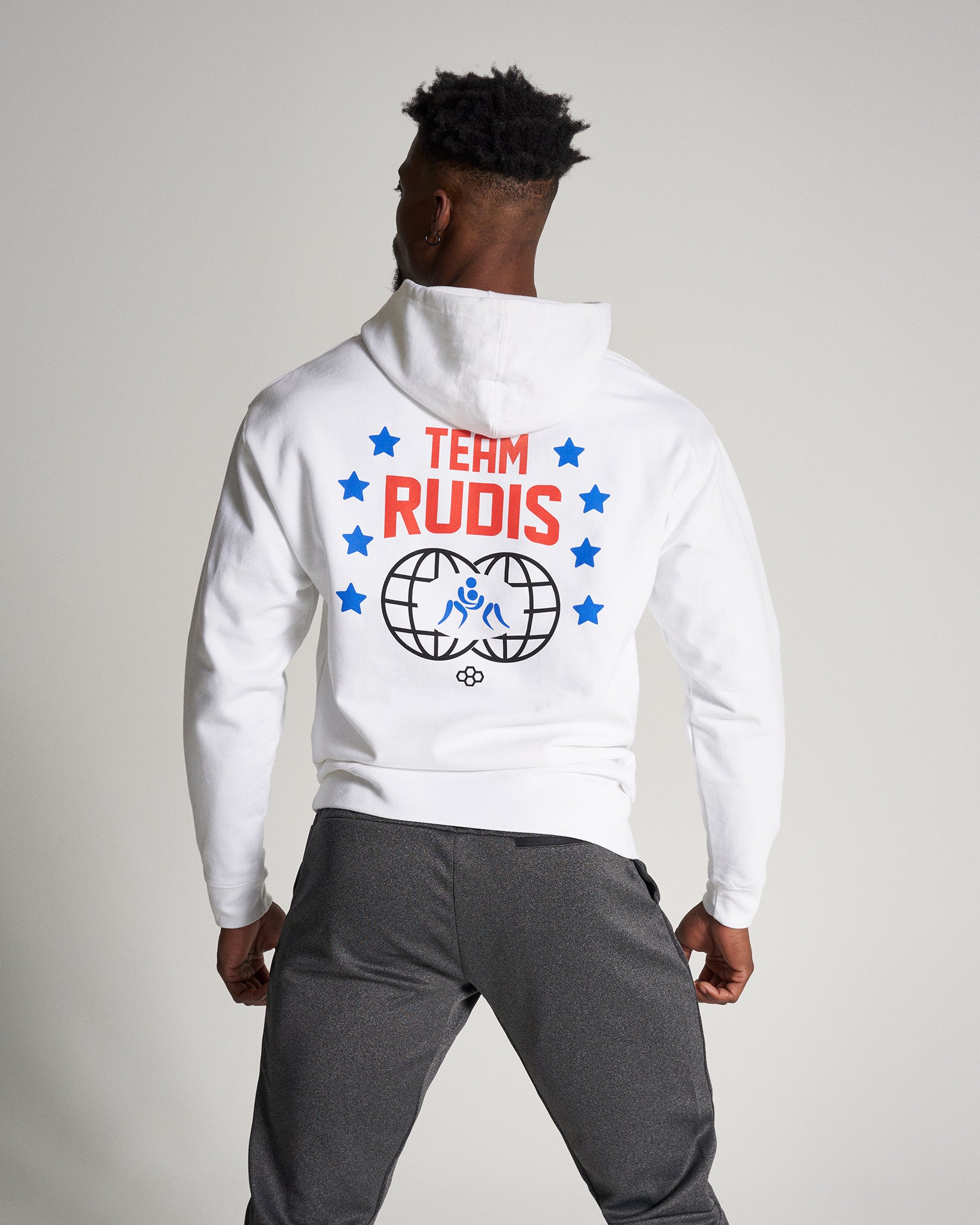 A man in a white hoodie with TEAM RUDIS printed on the back featuring a graphic design of a globe and stars paired with gray athletic pants