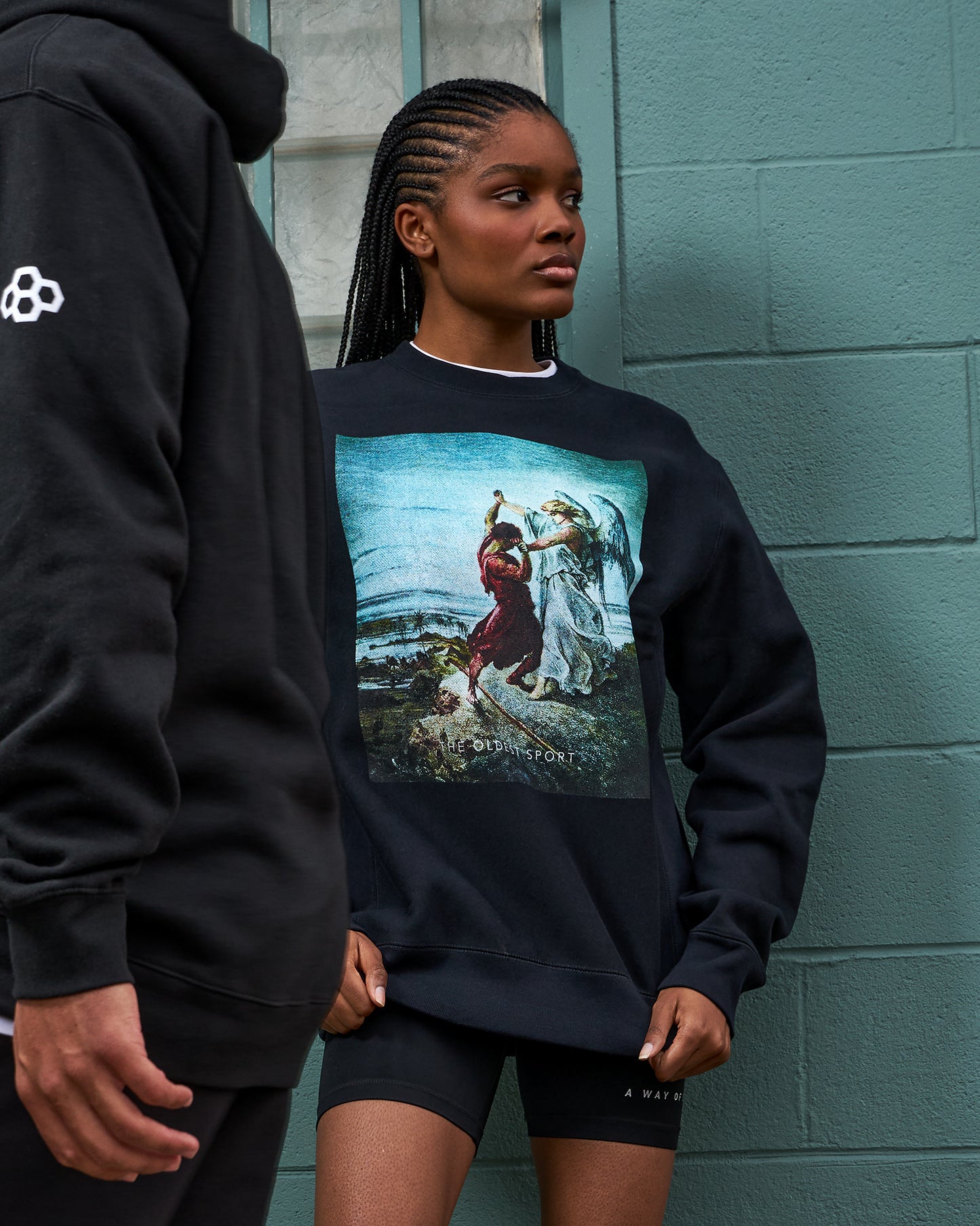 Person wearing a black sweatshirt with a graphic design, standing against a teal brick wall.