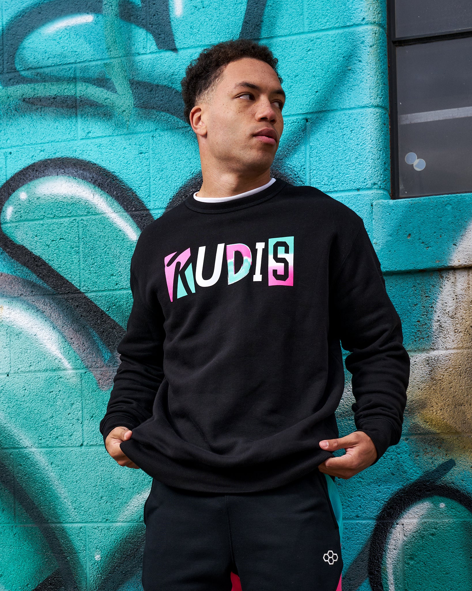 Person wearing a black sweatshirt with colorful text in front of a graffiti-covered wall.