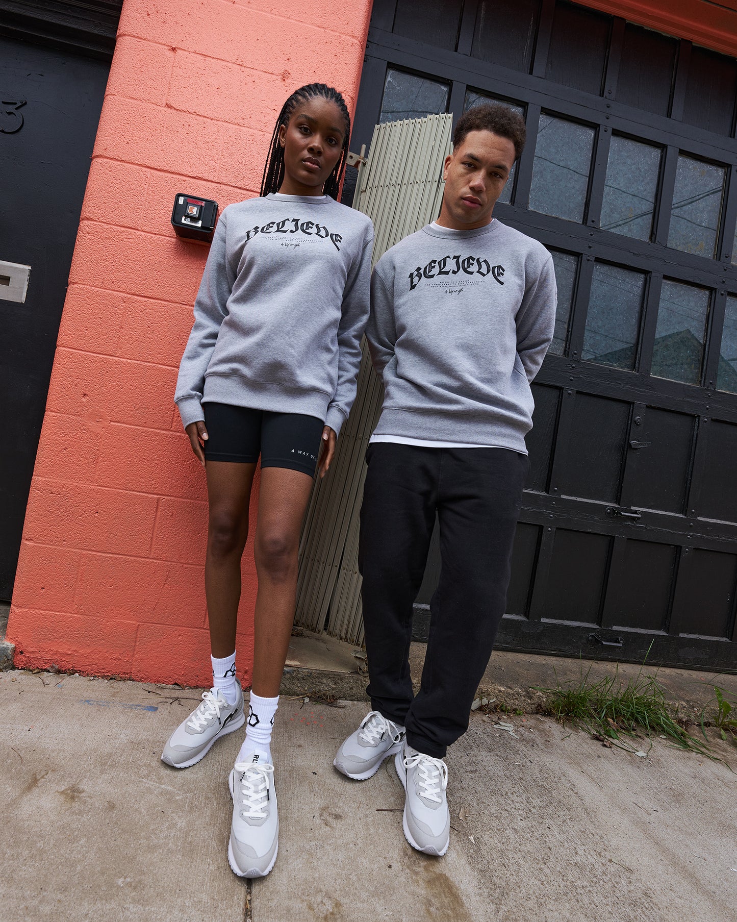 Two people wearing gray sweatshirts with 'believe' printed on them, standing in front of a red brick wall.