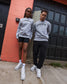 Two people wearing gray sweatshirts with 'believe' printed on them, standing in front of a red brick wall.