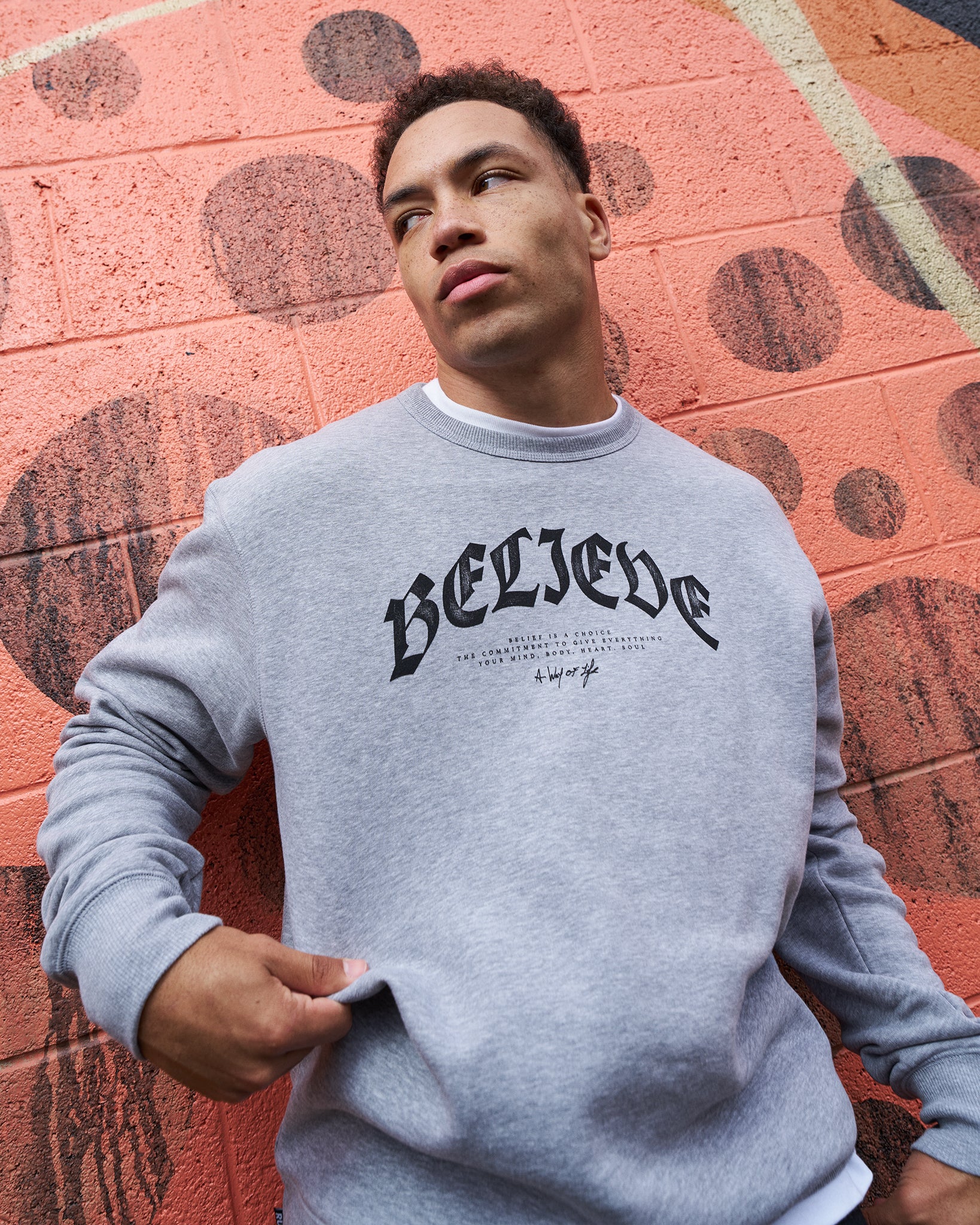Person wearing a gray sweatshirt with 'BELIEVE' text against a red and orange wall.